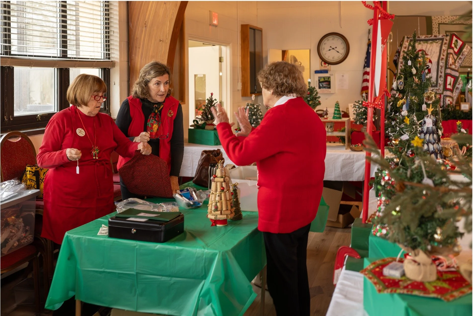 Three women at a Christmas craft sale table surrounded by decorated holiday trees and handmade ornaments