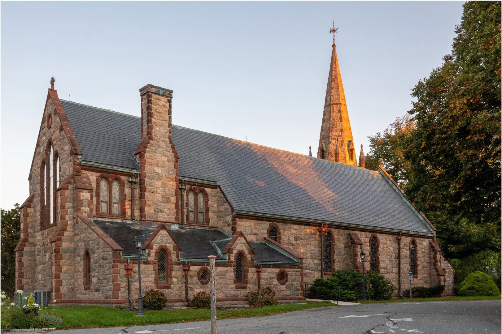 Historic Gothic Revival stone church with stained-glass windows, slate roof, and tall pointed steeple beside a tree-lined street