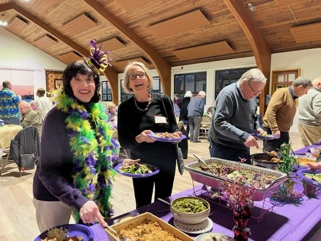 Guests wearing purple and green Mardi Gras accessories serve food at a festive buffet.