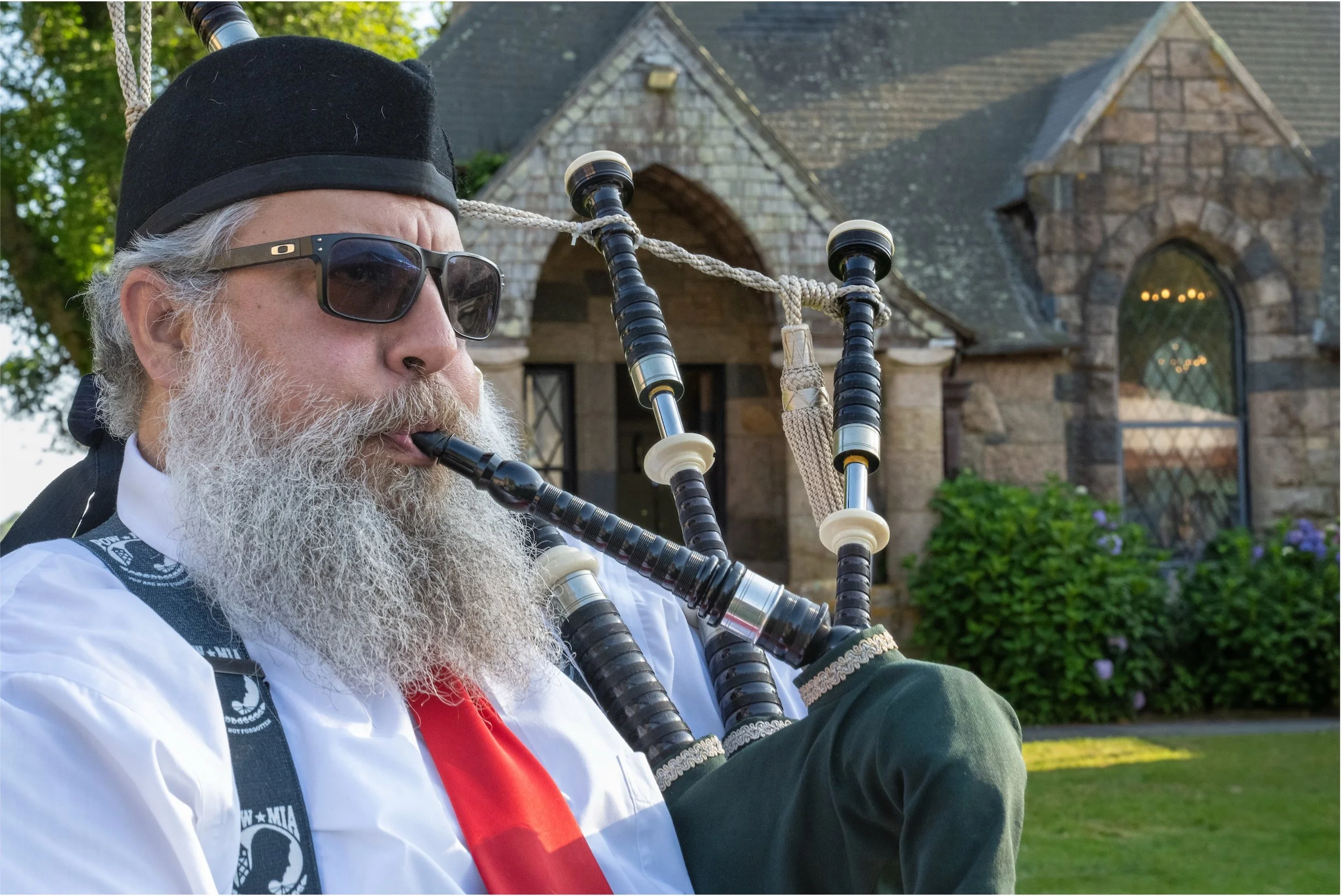 Bearded bagpiper wearing sunglasses and a black hat performs outside a stone church.