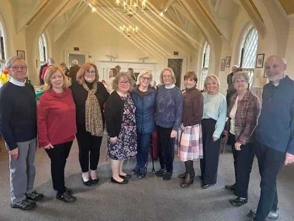 Group of eleven adults posing inside a bright Gothic-style church hall with chandeliers and stained-glass windows.