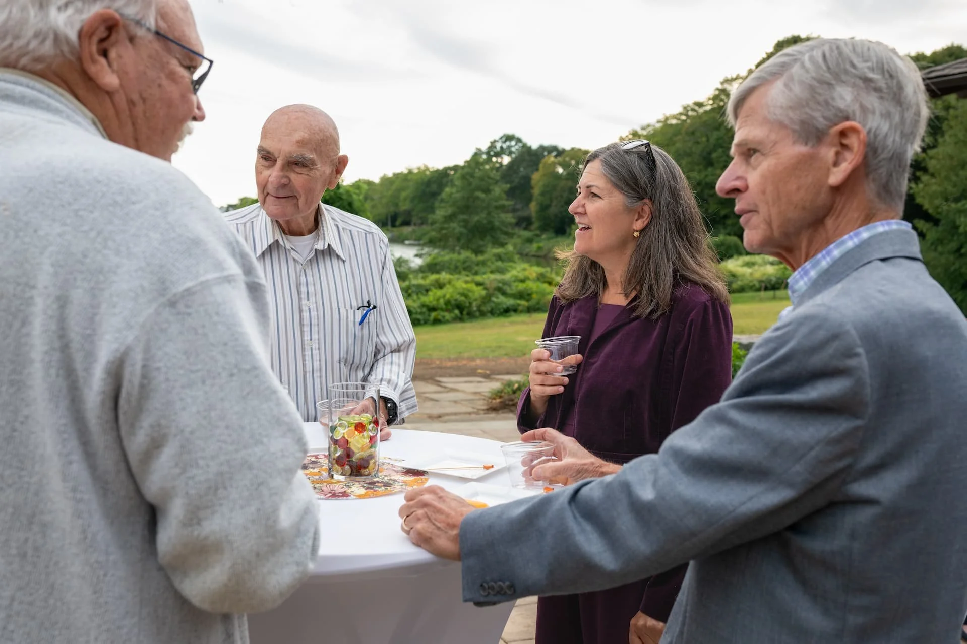 Four older adults socialize around an outdoor table with drinks in a garden setting