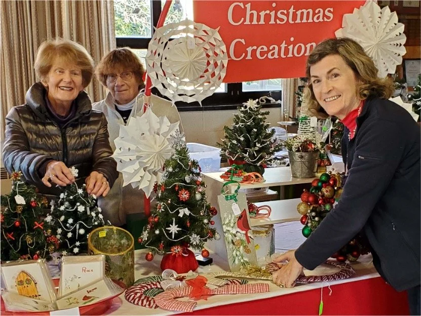 Three women display handmade Christmas decorations, miniature trees, wreaths, and holiday crafts at a Christmas creation event.