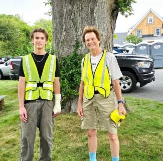 Two community volunteers wearing bright yellow reflective safety vests stand beside a large tree near parked vehicles and a house.