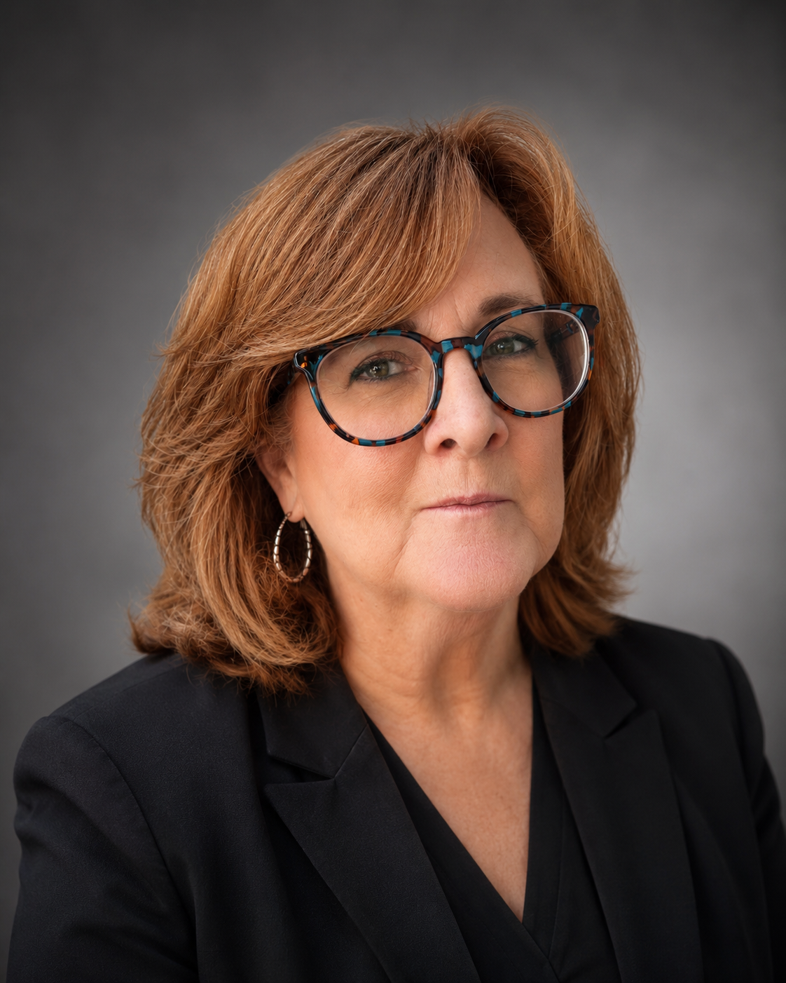 Professional headshot of a middle-aged woman wearing patterned eyeglasses and a black blazer against a gray background