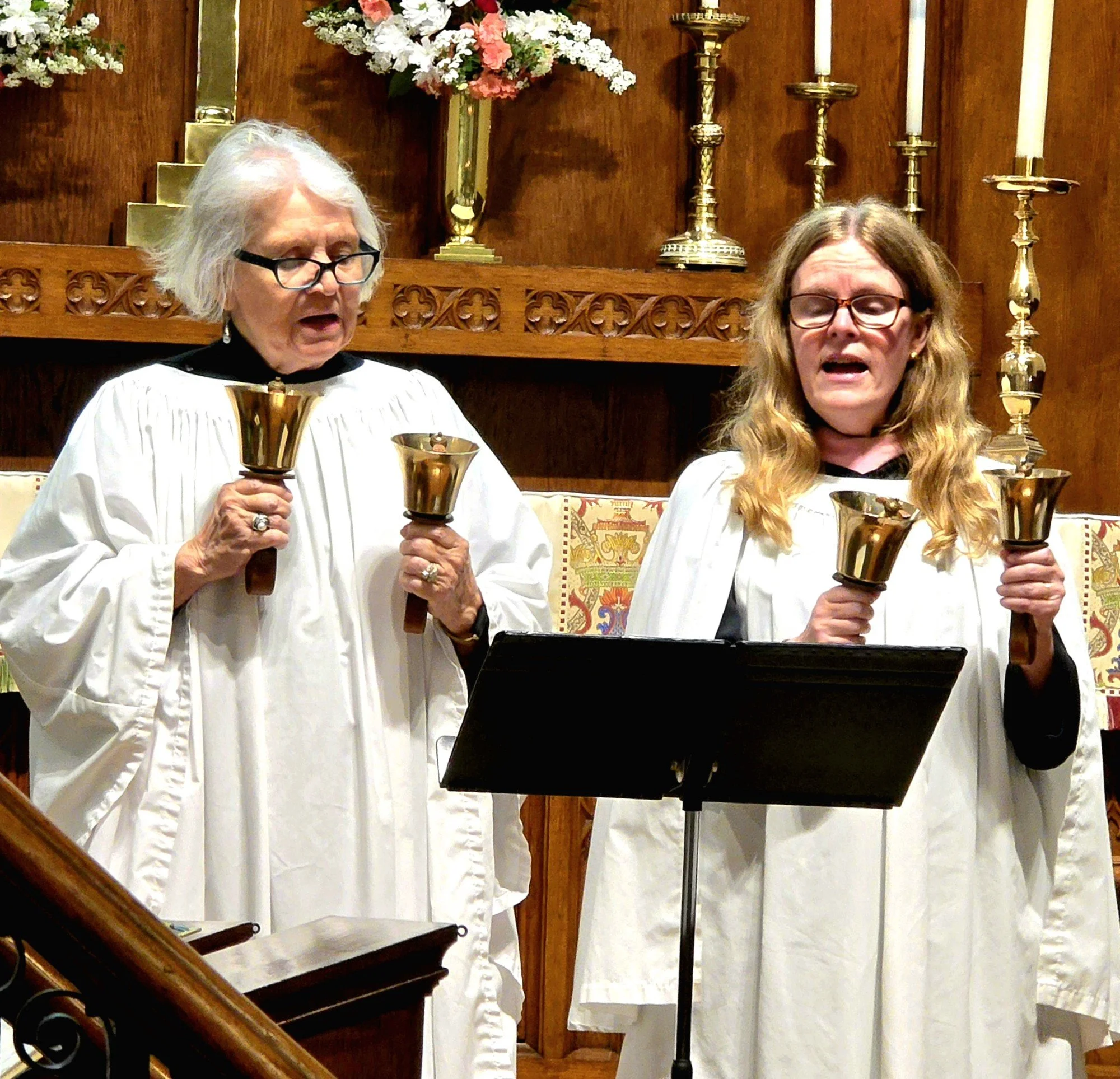 Two women in white choir robes sing while holding brass handbells inside a church.
