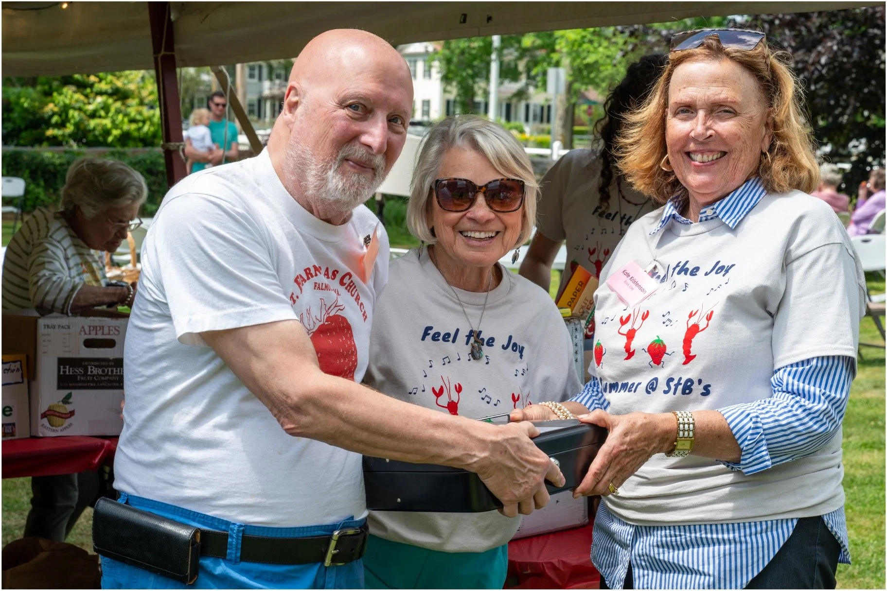 Three smiling adults pose at an outdoor summer event, holding a black case beneath a canopy.