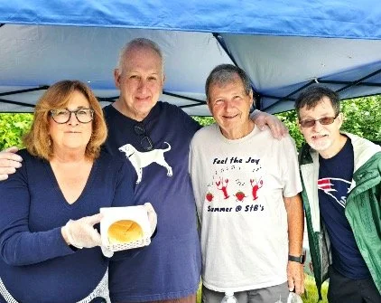 Four people pose under a blue canopy, with a woman holding a hamburger in a takeout container.