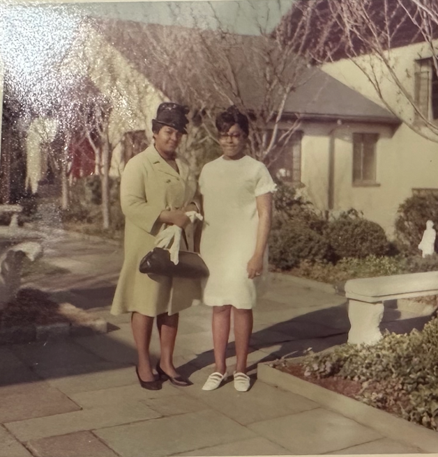 Vintage color photograph of two women standing on a residential sidewalk, one wearing a beige coat and hat and the other in a white dress and sneakers.