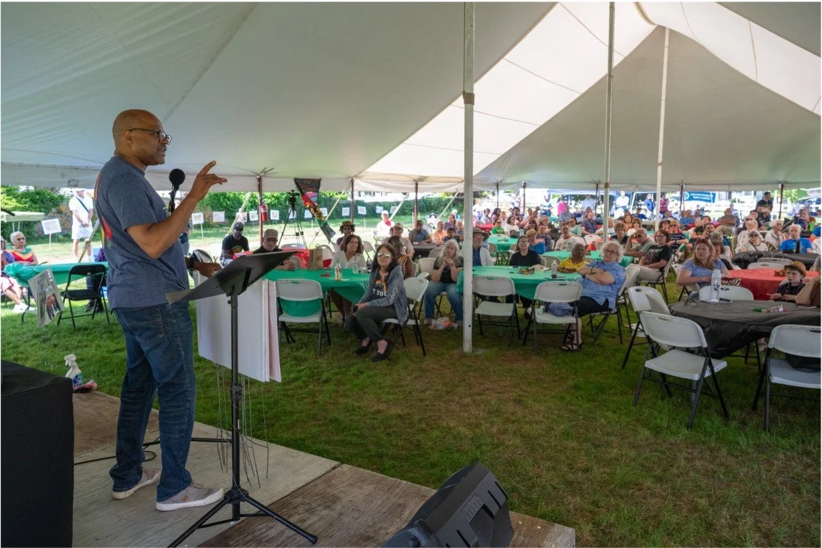 Speaker addressing a seated audience beneath a large outdoor event tent.