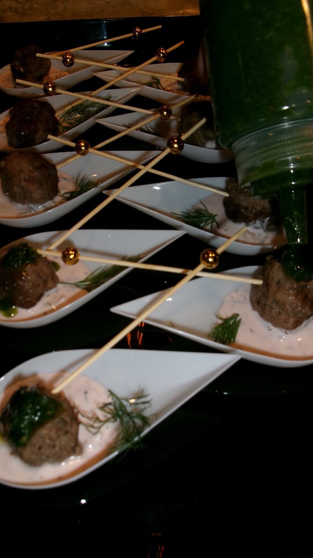 Appetizers of meatballs served in small white dishes with sauce and garnished with herbs, arranged on a dark surface with a green bottle nearby.