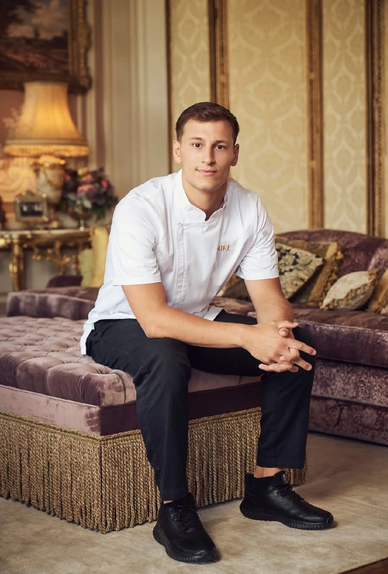 A young man in a white chef's coat sitting on a plush, vintage-style sofa in an elegant living room with classic wallpaper, a matching sofa set, and ornate gold accents.