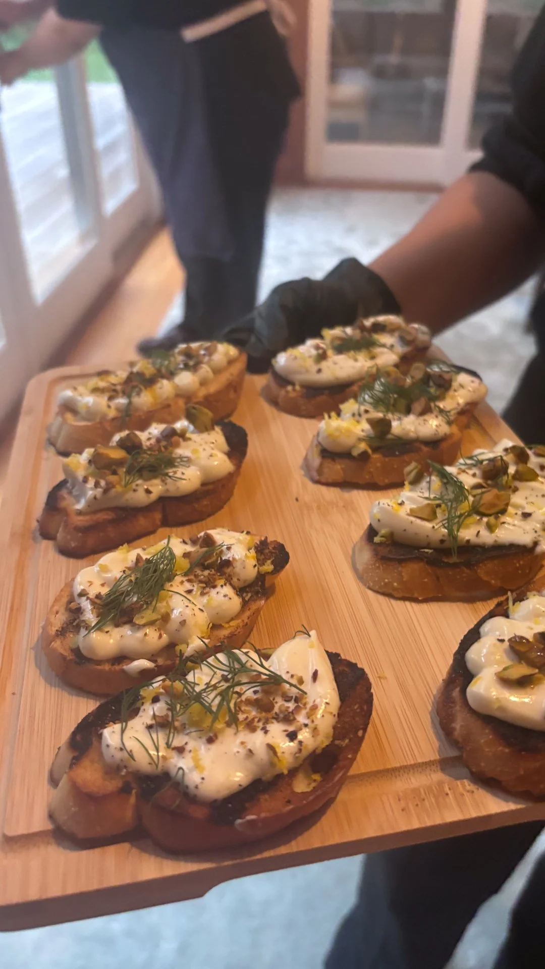 Chef Mike Catuosco - Gallery - Close-up of toasted bread slices topped with creamy white cheese, chopped nuts, herbs, and garnished with herbs on a wooden serving board.