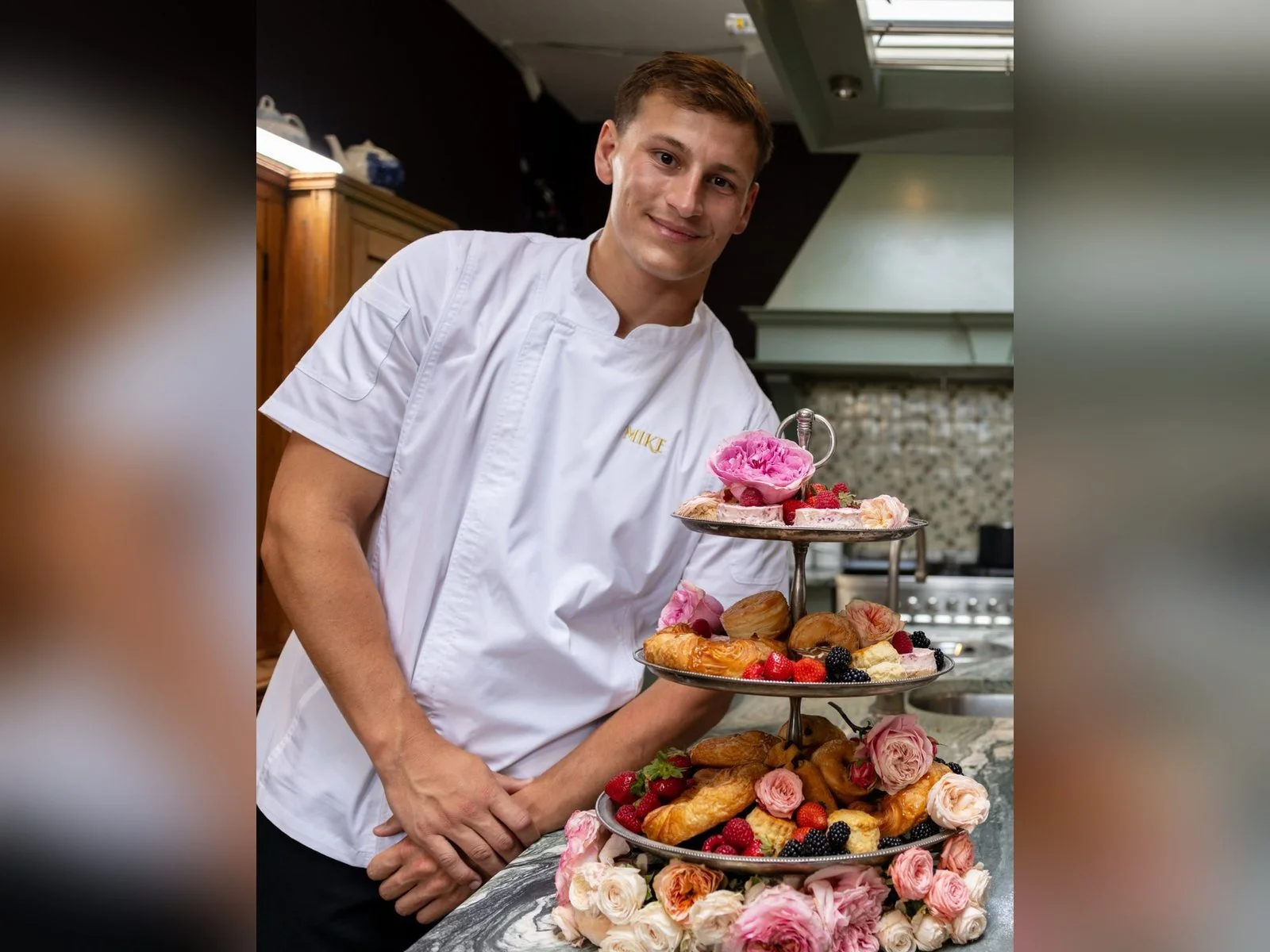 A young man in a white chef's jacket smiling, standing next to a multi-tiered tray filled with pastries, berries, and decorated with pink and white roses, in a kitchen setting.