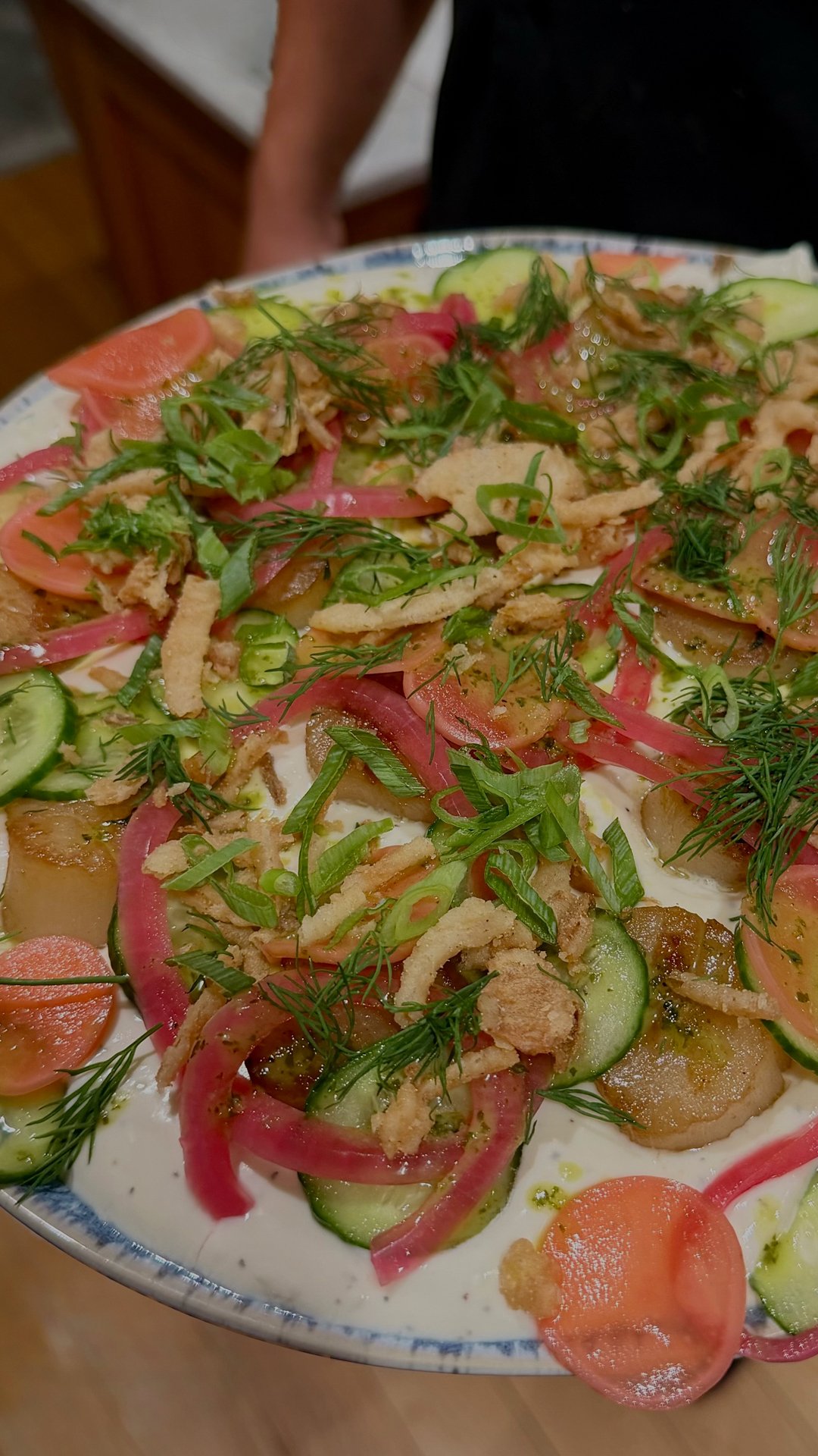 Close-up of a plate with salad containing sliced cucumbers, tomatoes, pickled onions, fresh herbs, and crispy fried elements, possibly breaded fish or chicken strips.