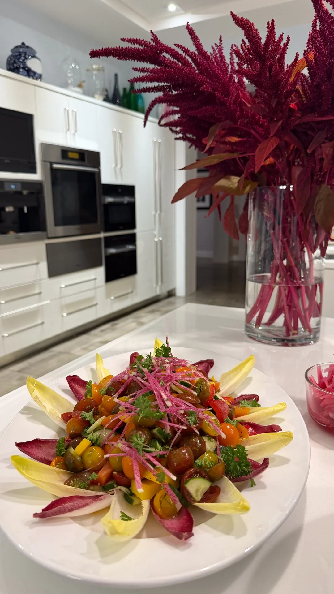 Chef Mike Catuosco - Gallery - A colorful vegetable salad garnished with herbs on a white plate, with a large pinkish-red floral arrangement in a clear glass vase in the background on a kitchen counter.