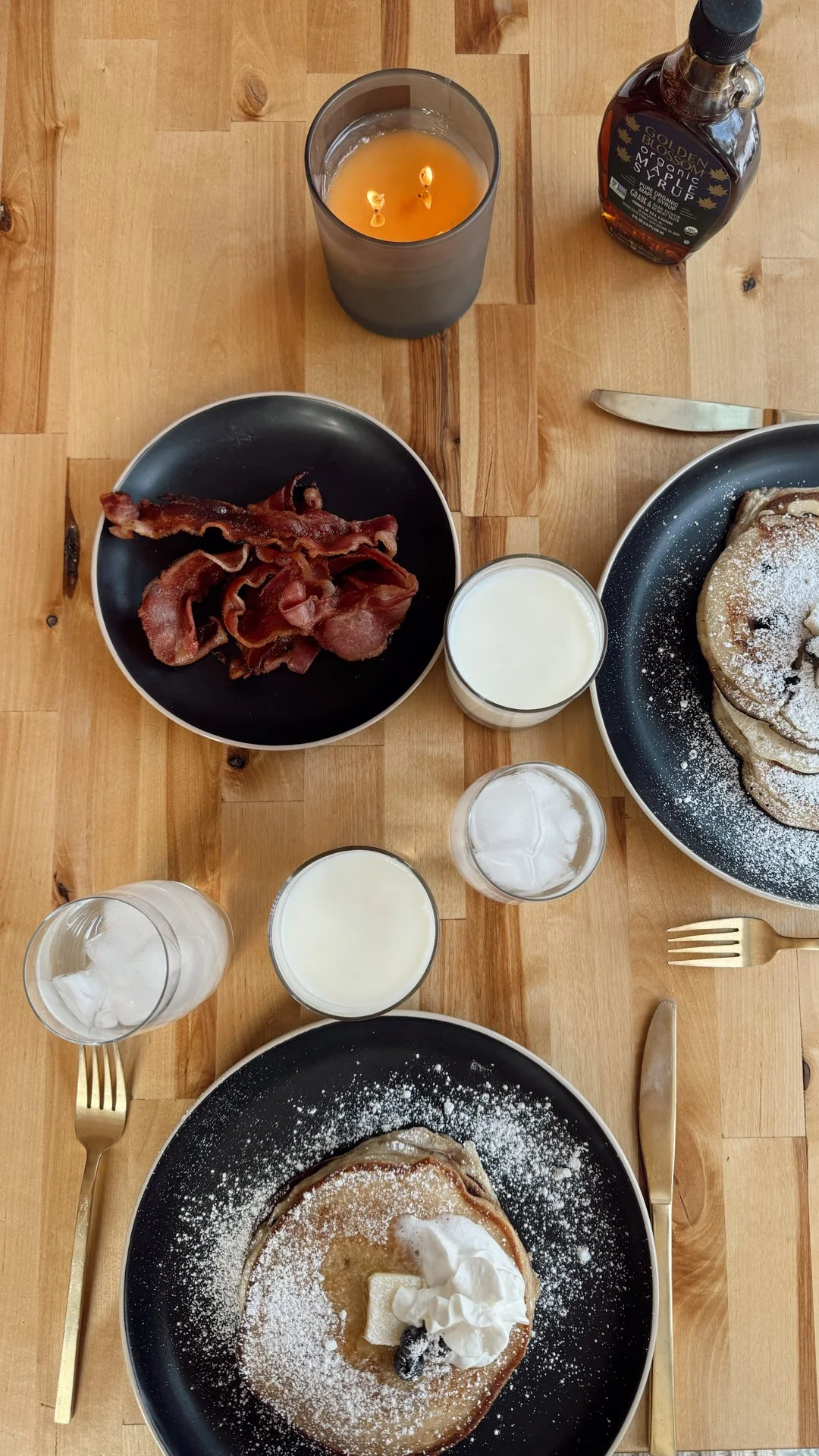 Chef Mike Catuosco - Gallery - Breakfast scene with two plates of pancakes dusted with powdered sugar, whipped cream, and fruit, two glasses of milk with ice, a bowl of crispy bacon, a lit candle, and a bottle of syrup on a wooden table.