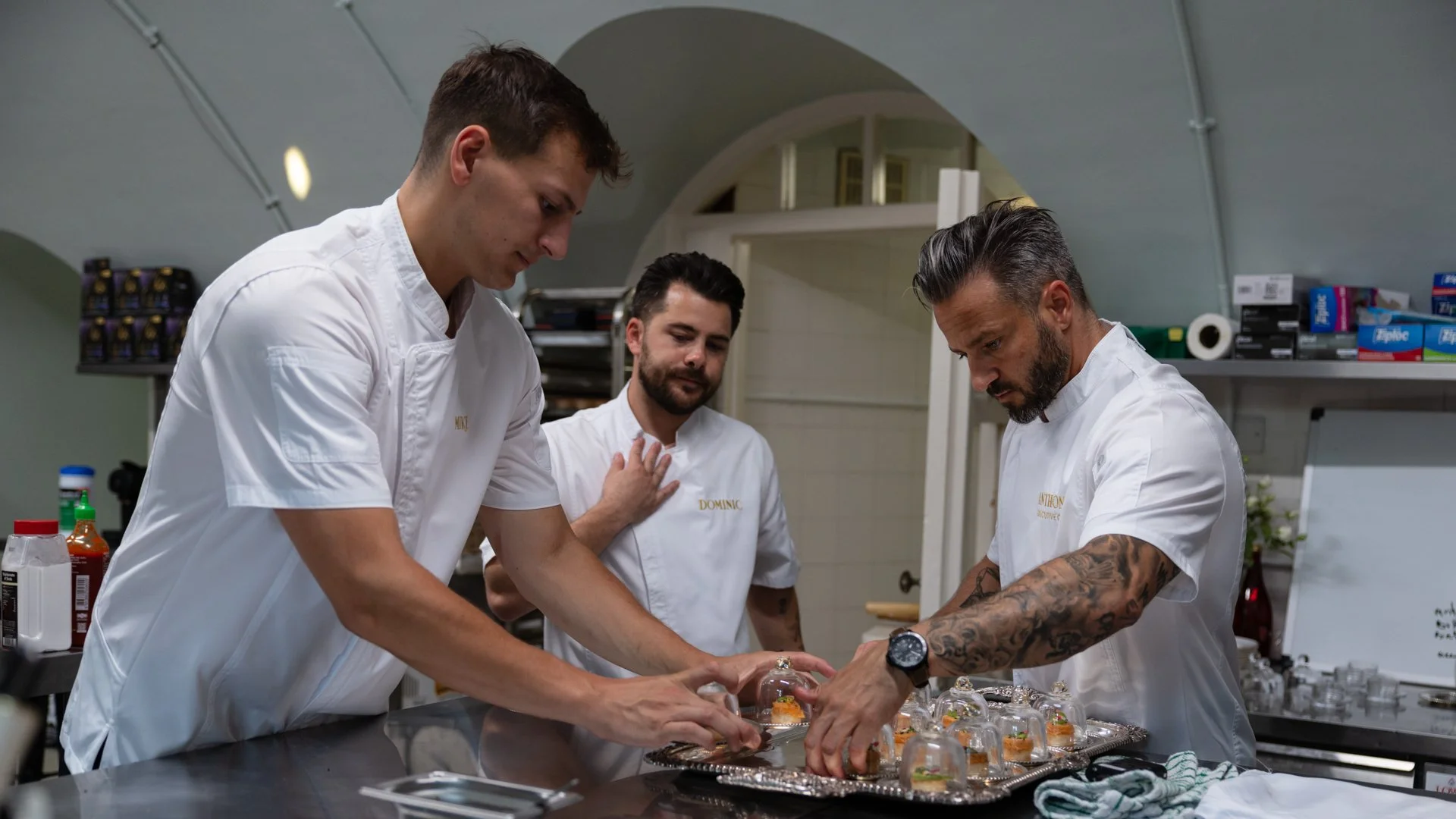 Three chefs in white uniforms preparing plated dishes in a kitchen.