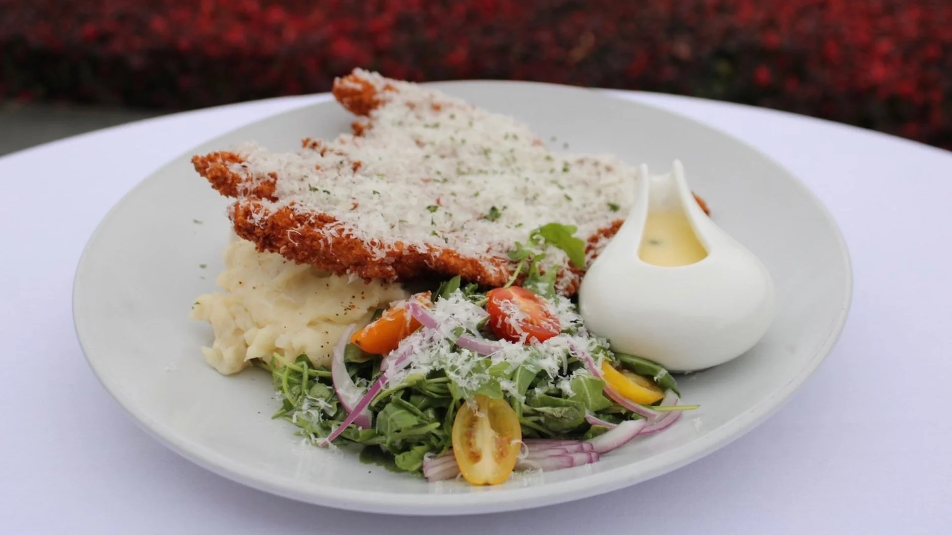 Plate with breaded fried cutlet topped with grated cheese, served with potatoes, fresh salad with cherry tomatoes, sliced red onions, and sprinkled with grated cheese, alongside a dollop of white sauce or dressing.