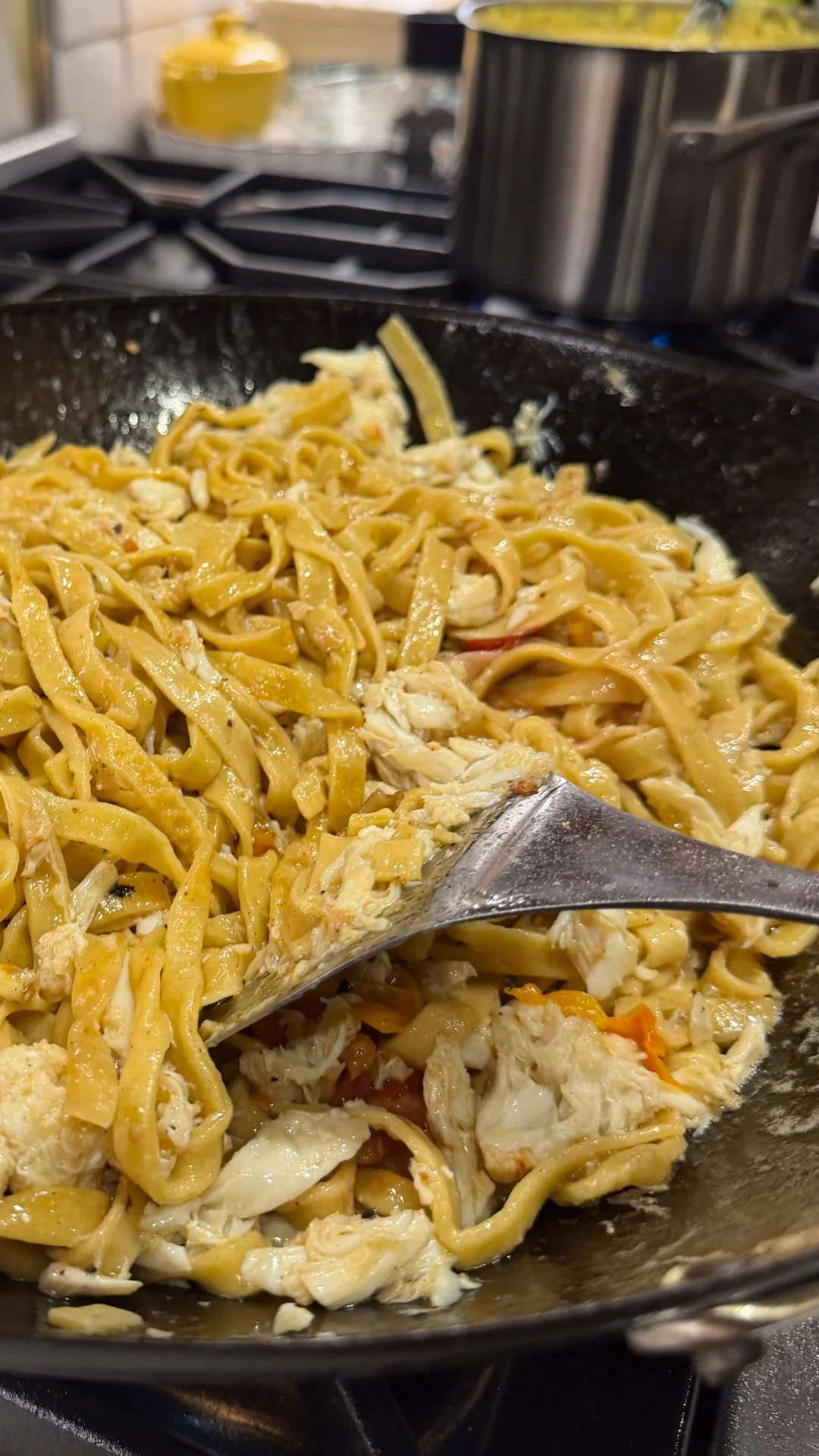 Chef Mike Catuosco - Gallery - Close-up of cooked chicken and pasta with sauce in a black skillet on a stovetop, with a metal spatula stirring the dish.