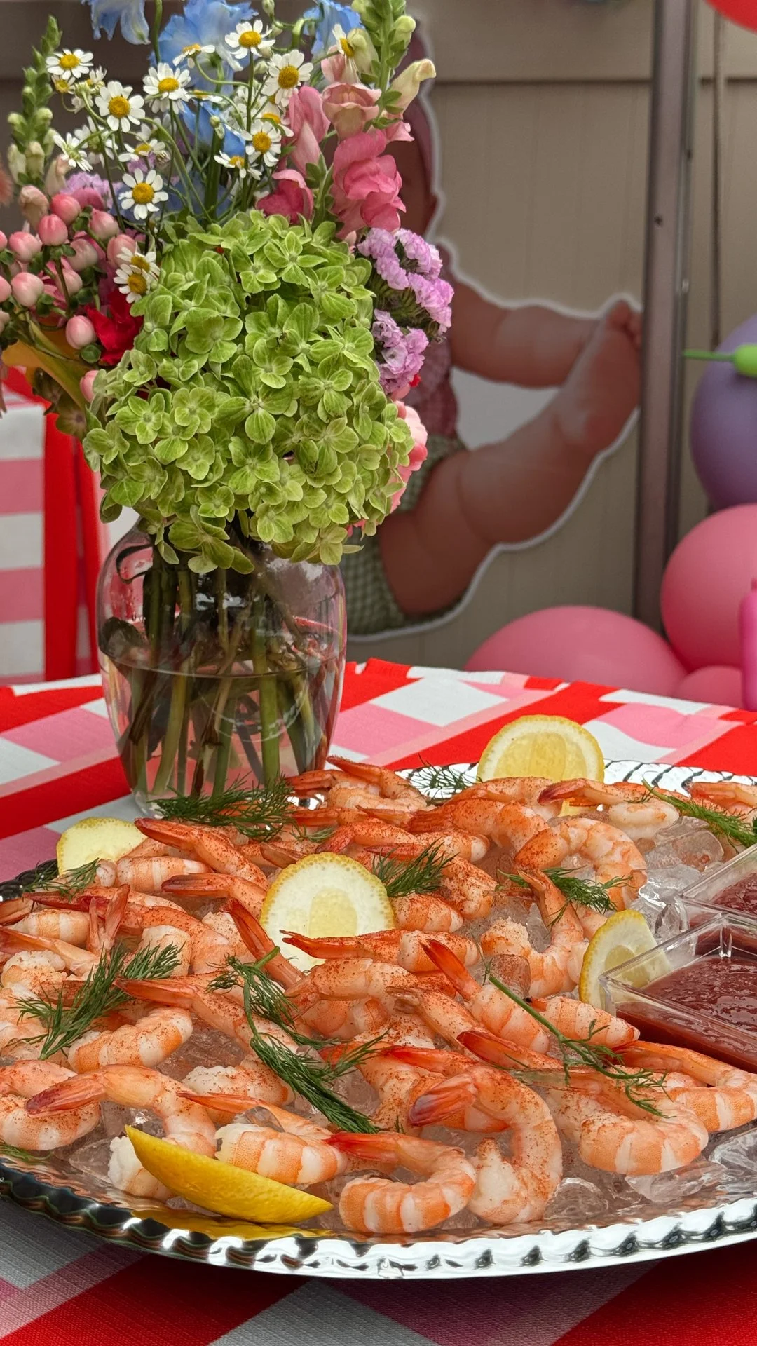 A table with a platter of cooked shrimp garnished with lemon slices and fresh dill, set on a red and white checkered tablecloth, with a large bouquet of colorful flowers in a glass vase in the background.