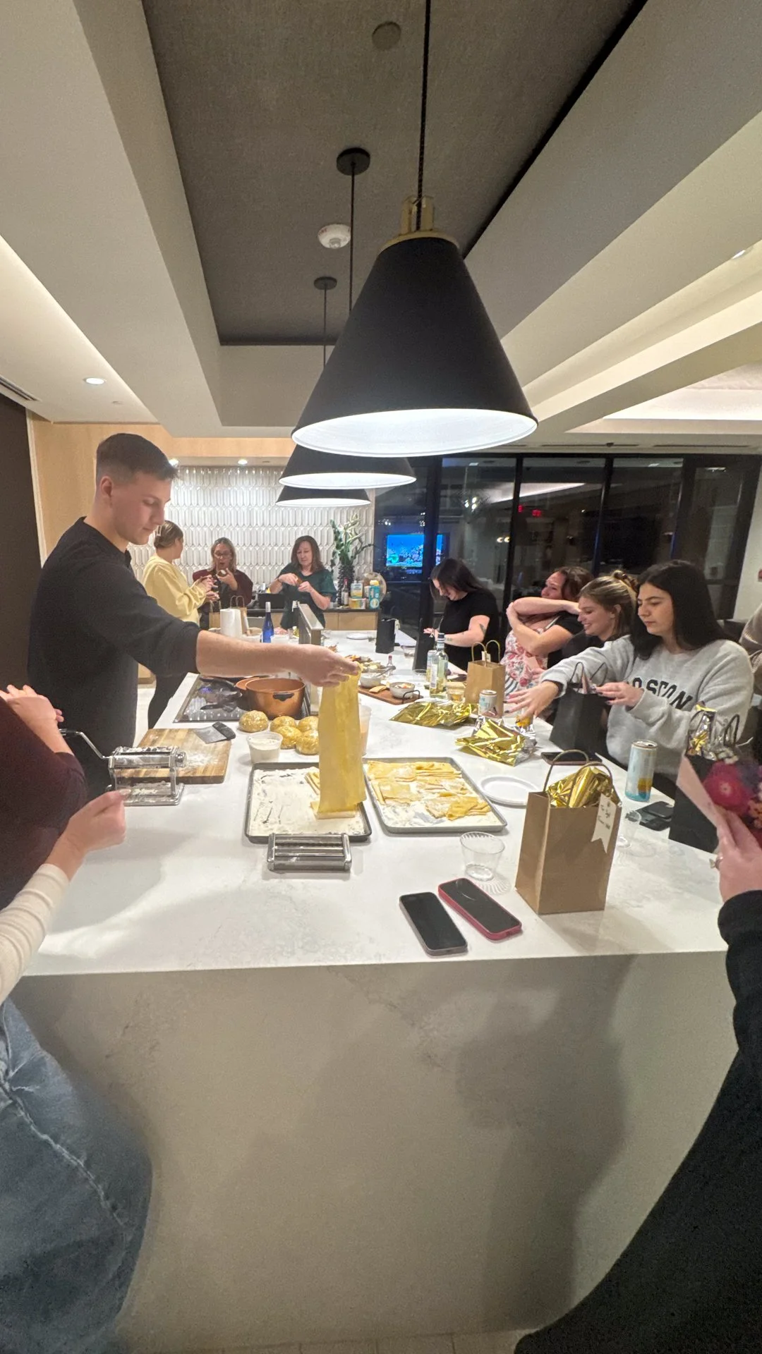 People gathered around a kitchen island, decorating cookies and wrapping presents during a holiday celebration. A young man is serving food or snacks, with gift bags and food on the counter.