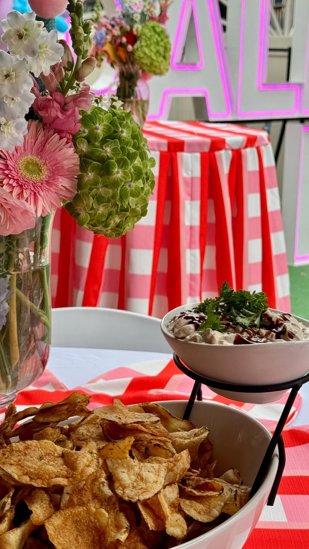Chef Mike Catuosco - Gallery - A table with a bowl of potato chips, a bowl of cream dip topped with herbs and sauce, a flower arrangement in a glass vase, and another floral arrangement in the background.