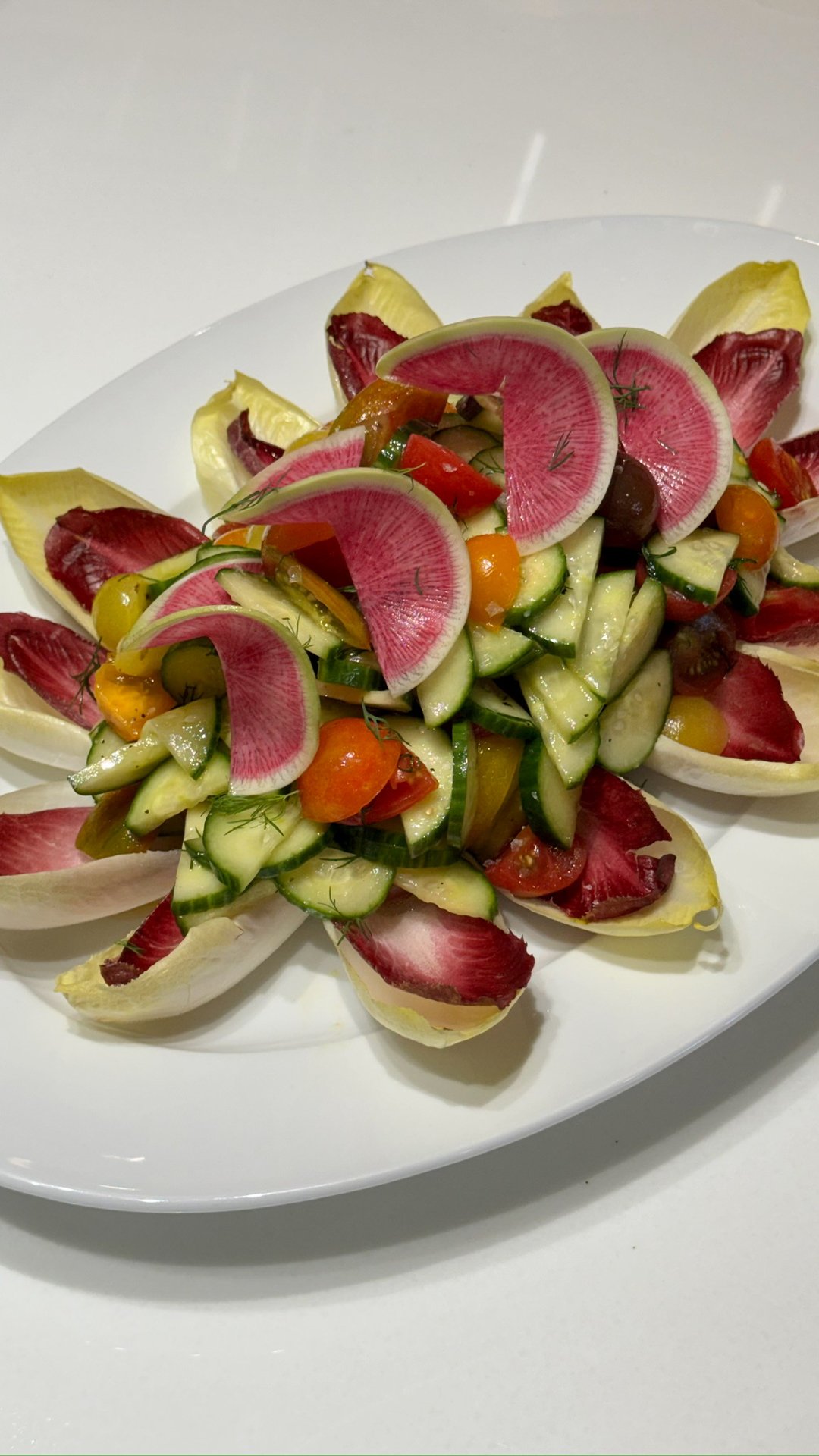 A colorful vegetable salad arranged on a white oval platter, featuring sliced cucumbers, cherry tomatoes, radish slices, grapes, and endive leaves.