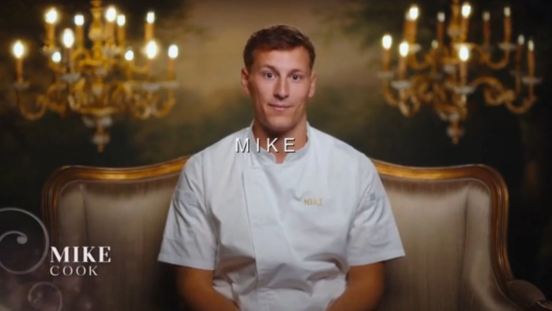A man named Mike sitting on a vintage-style sofa in a room with dark walls and ornate chandeliers, wearing a white chef's coat with his name embroidered on it.