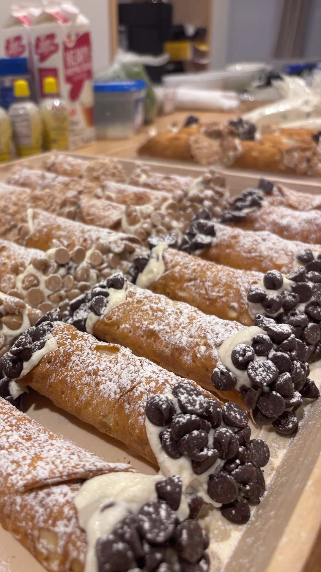 Row of cannoli filled with cream and topped with chocolate chips in a bakery or kitchen setting.