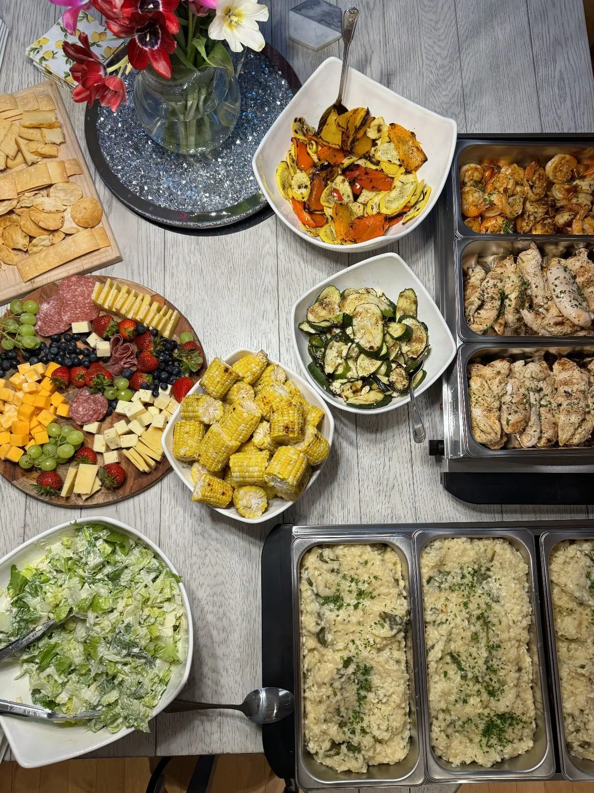 Table with various food dishes including roasted vegetables, grilled chicken, cheese platter, corn on the cob, potato salad, and a vegetable casserole, topped with a vase of flowers.