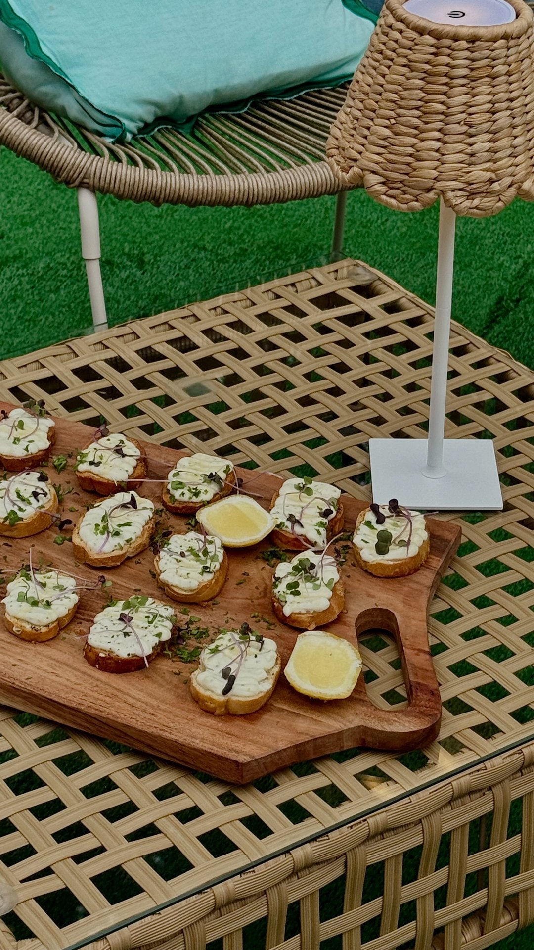 Wooden platter with toasted bread slices topped with creamy spread, microgreens, and herbs, garnished with lemon wedges, on a bamboo table, with a wicker lamp and a cushion in the background.