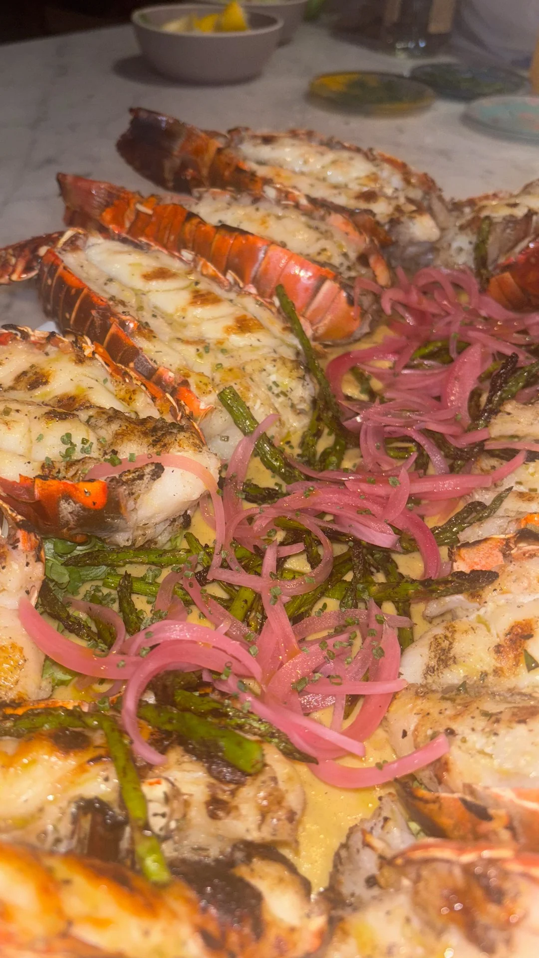 Close-up of grilled lobster tails topped with melted cheese, served with grilled asparagus and pickled red onions.