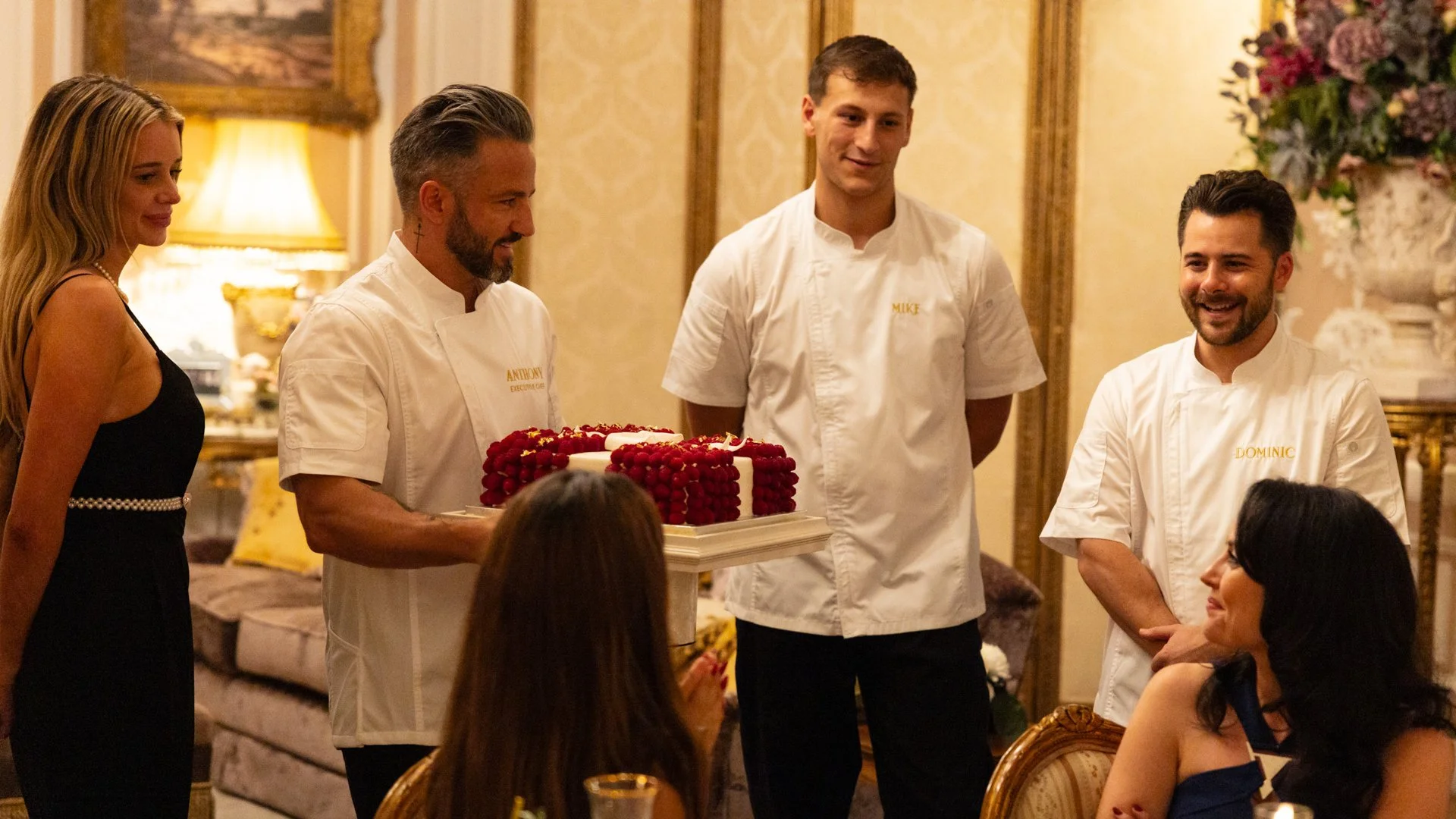 Three men in white chef jackets and two women in evening dresses at an elegant celebration in a luxurious room, with one man holding a cake decorated with red berries.