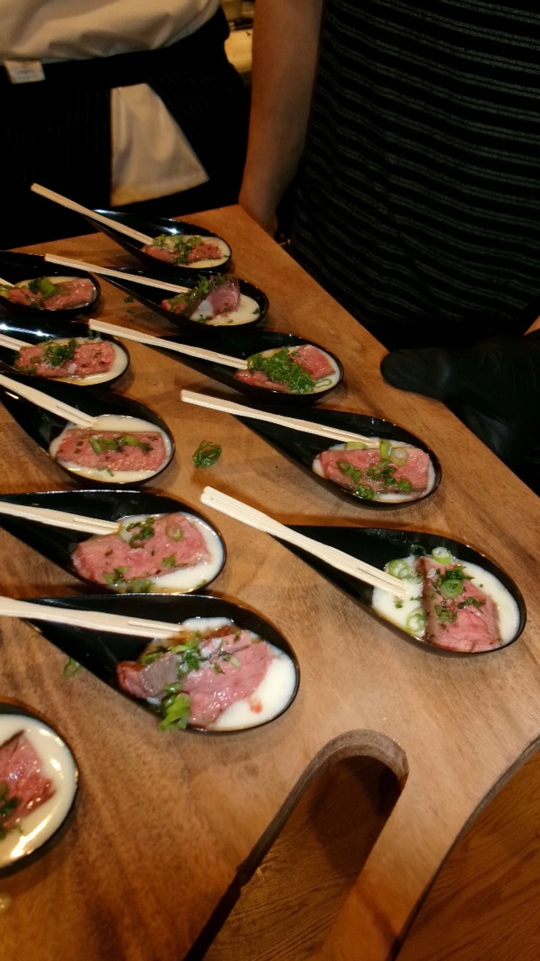 Sliced raw meat on spoons with green onions and sauce, placed on a wooden surface with chopsticks resting on each spoon.