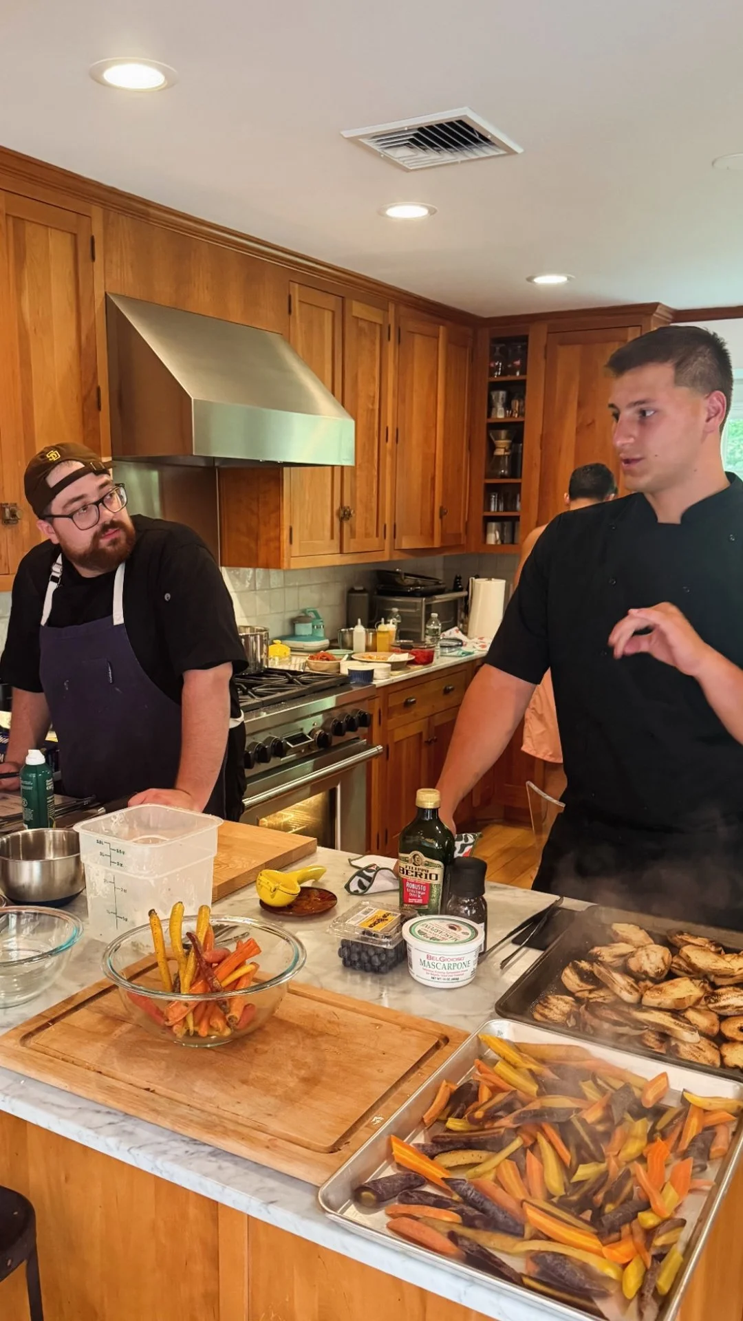 Chef Mike Catuosco - Gallery - Two chefs in a kitchen having a conversation during cooking. The countertop has roasted vegetables and various cooking ingredients and utensils. Wooden cabinets and a stainless steel range hood are in the background.