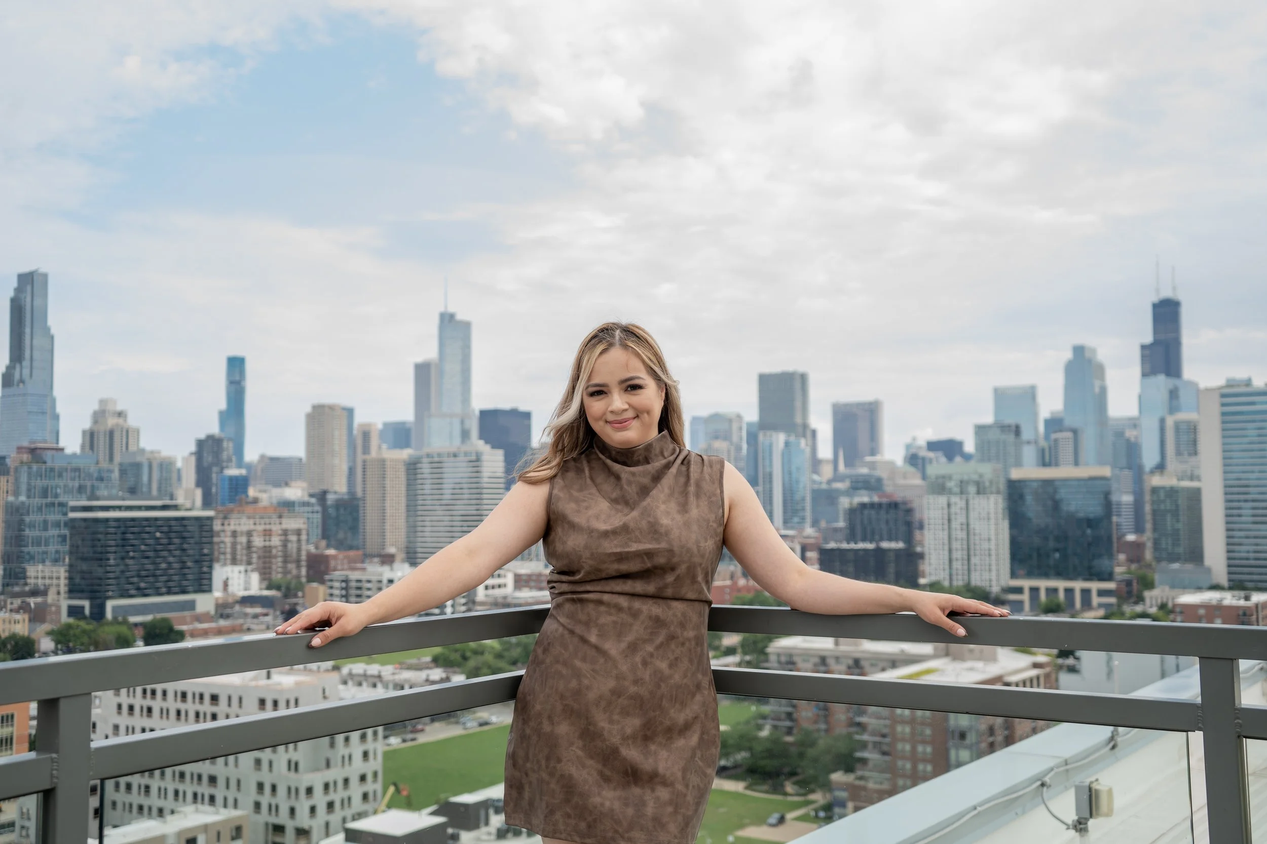 A woman with long hair stands on a rooftop with a city skyline behind her. She is smiling and leaning on a metal railing.