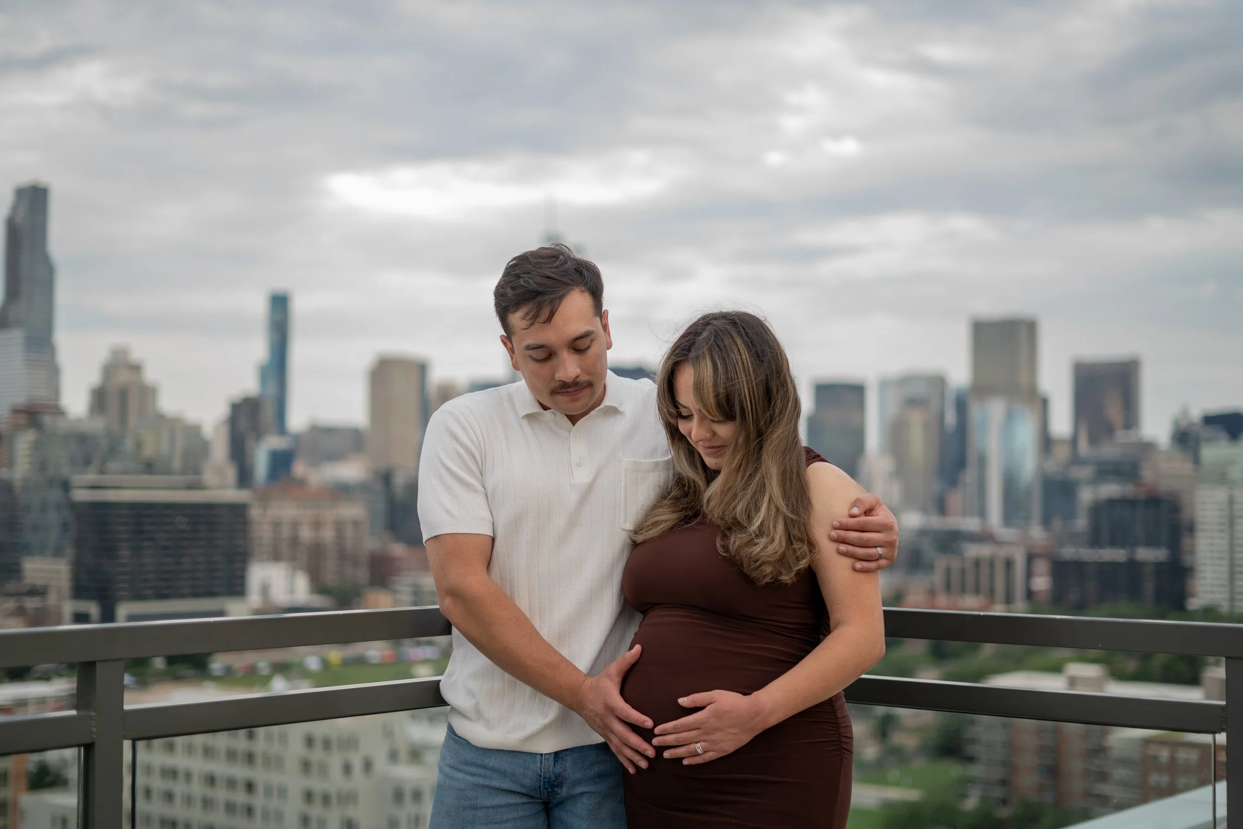 A man and pregnant woman standing close together on a balcony overlooking a city skyline on an overcast day, with the man holding the woman’s belly.
