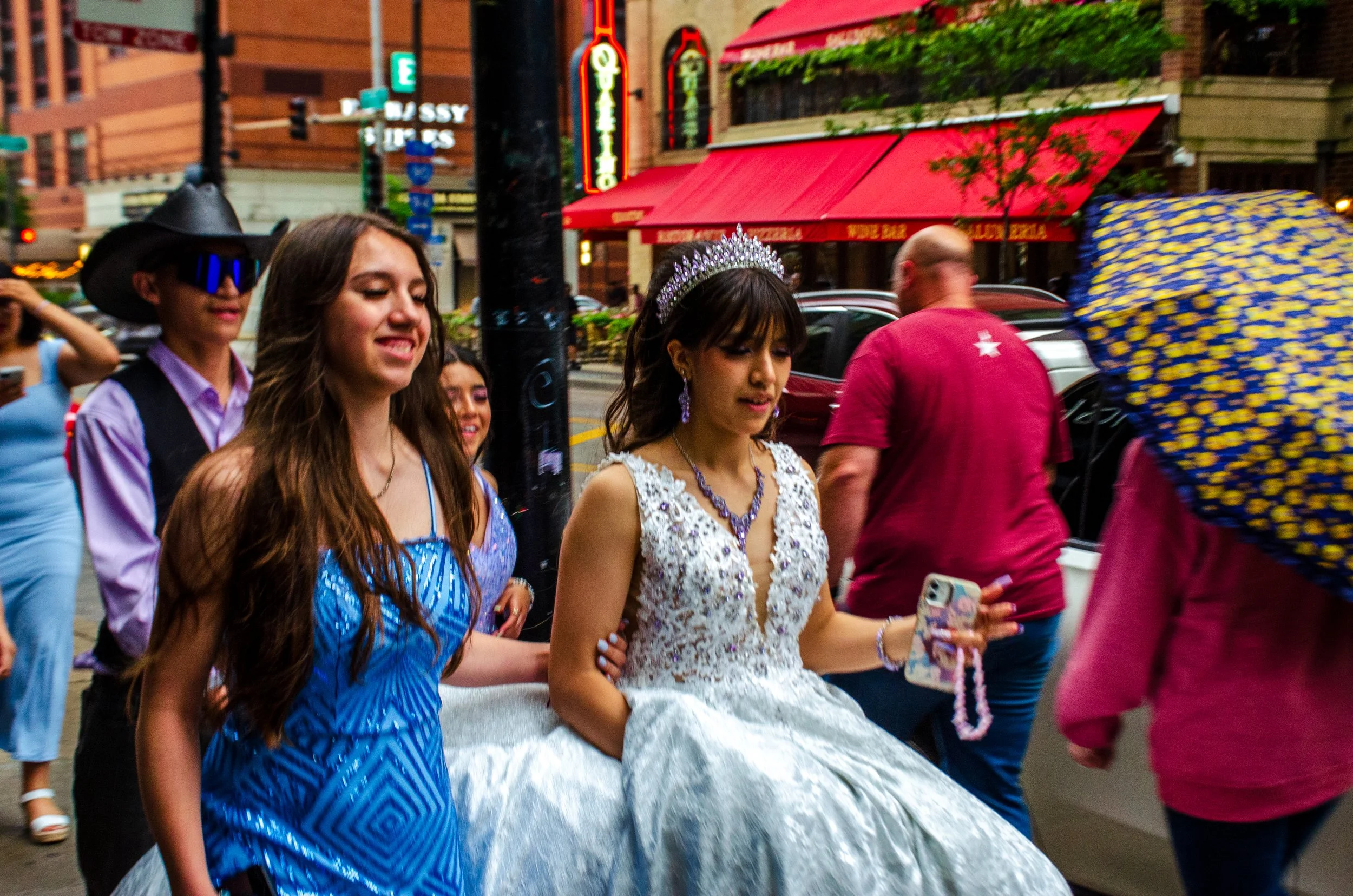 Young women dressed in fancy gowns walking outdoors, with some wearing tiaras and jewelry, in a parade or event setting on a city street