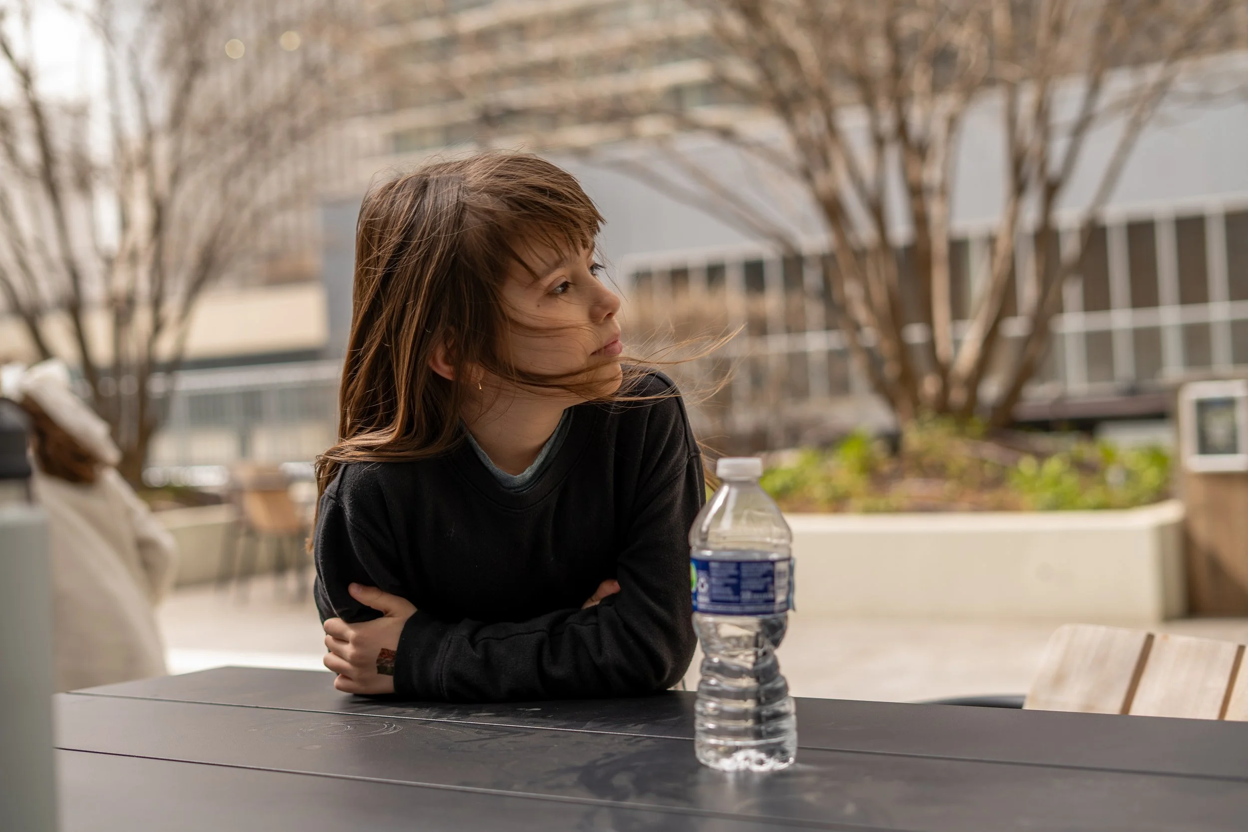 A young woman with brown hair, casual black sweater, sitting at a table outdoors, arms crossed, with a plastic water bottle in front of her. The background shows leafless trees and modern buildings, suggesting a chill, outdoor city setting.