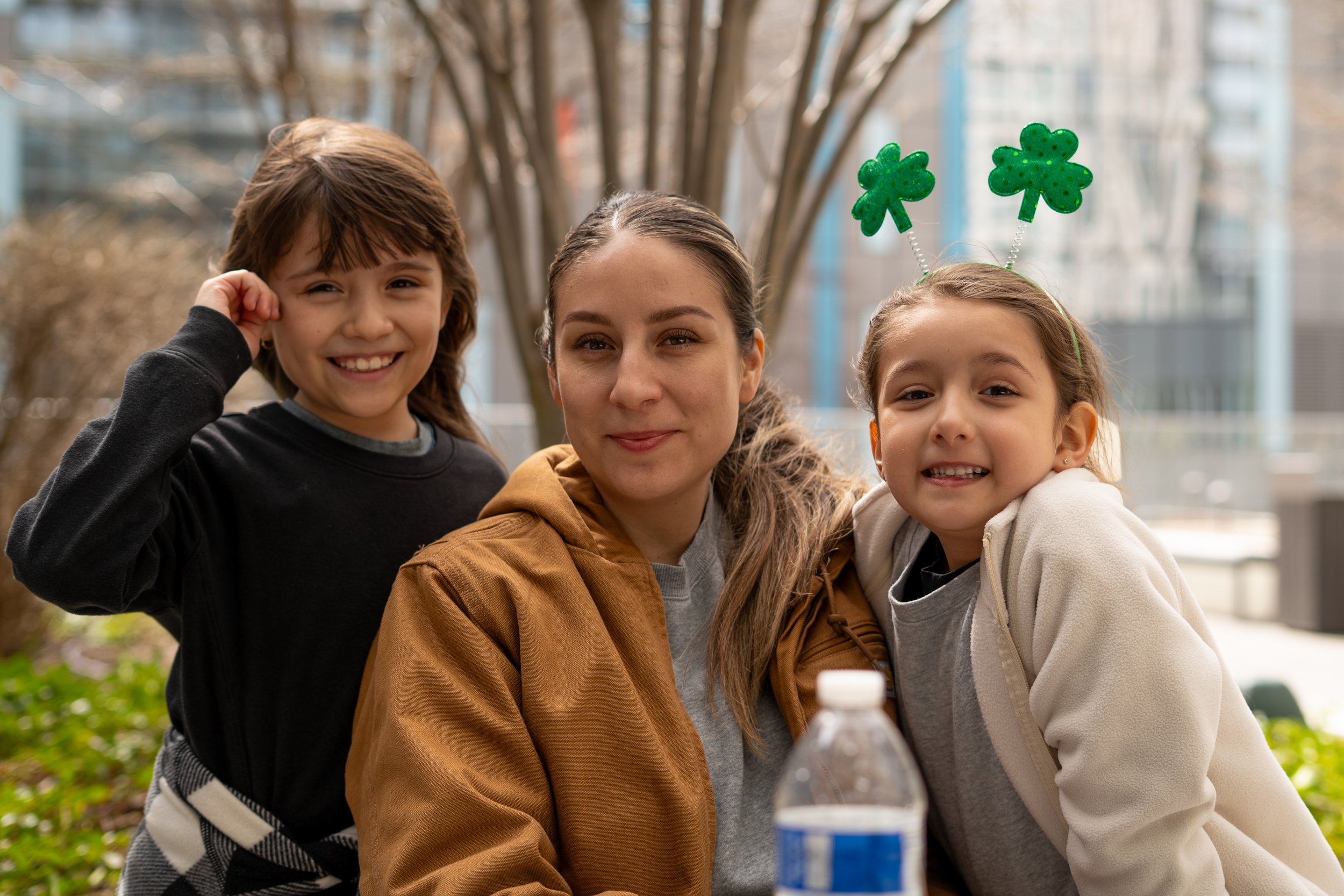 Three women smiling outdoors, one wearing a St. Patrick's Day headband with shamrocks, with trees and buildings in the background.