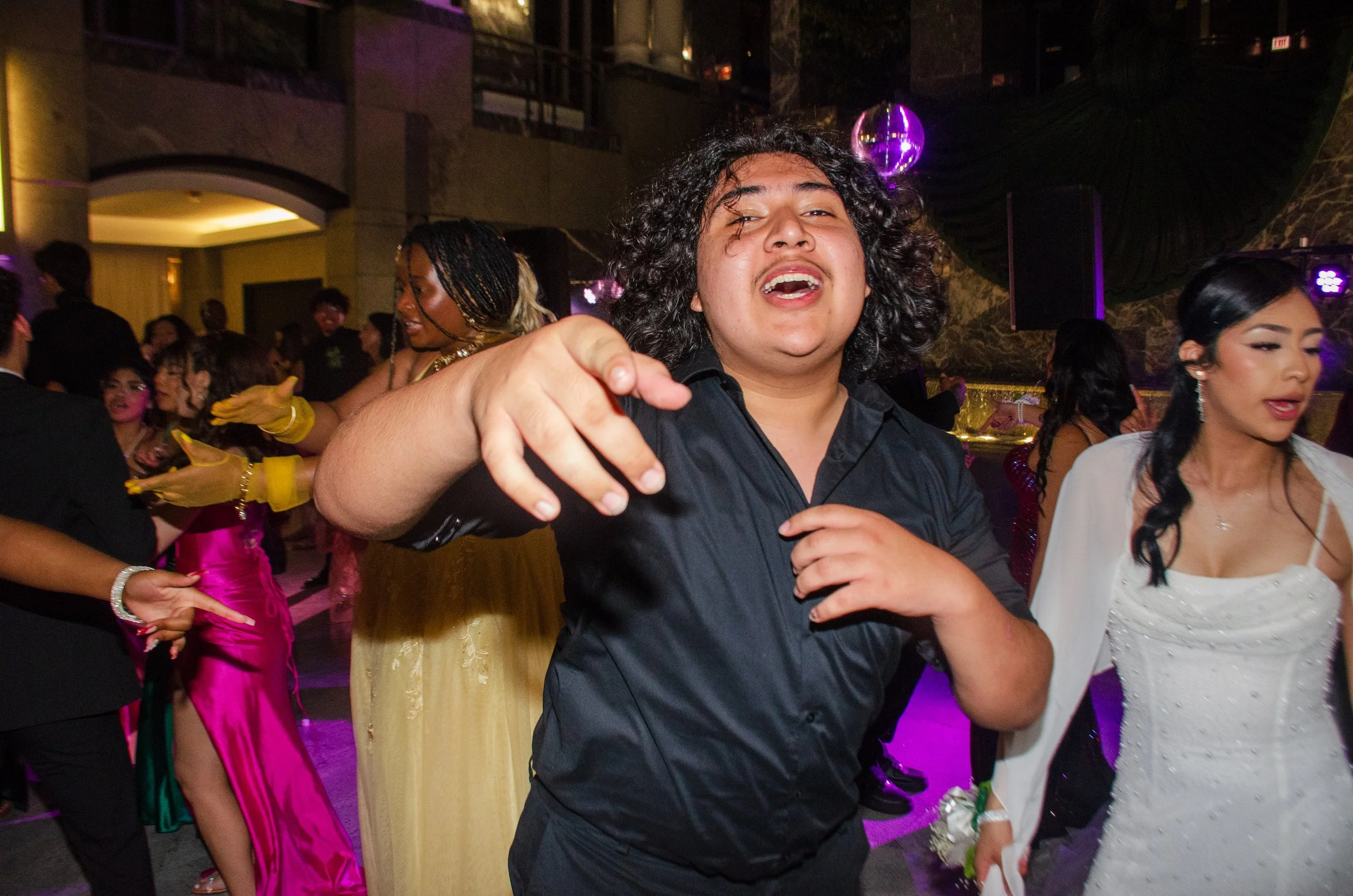 People dancing and having fun at a lively indoor party, with a man in the foreground pointing towards the camera, surrounded by women in colorful dresses.