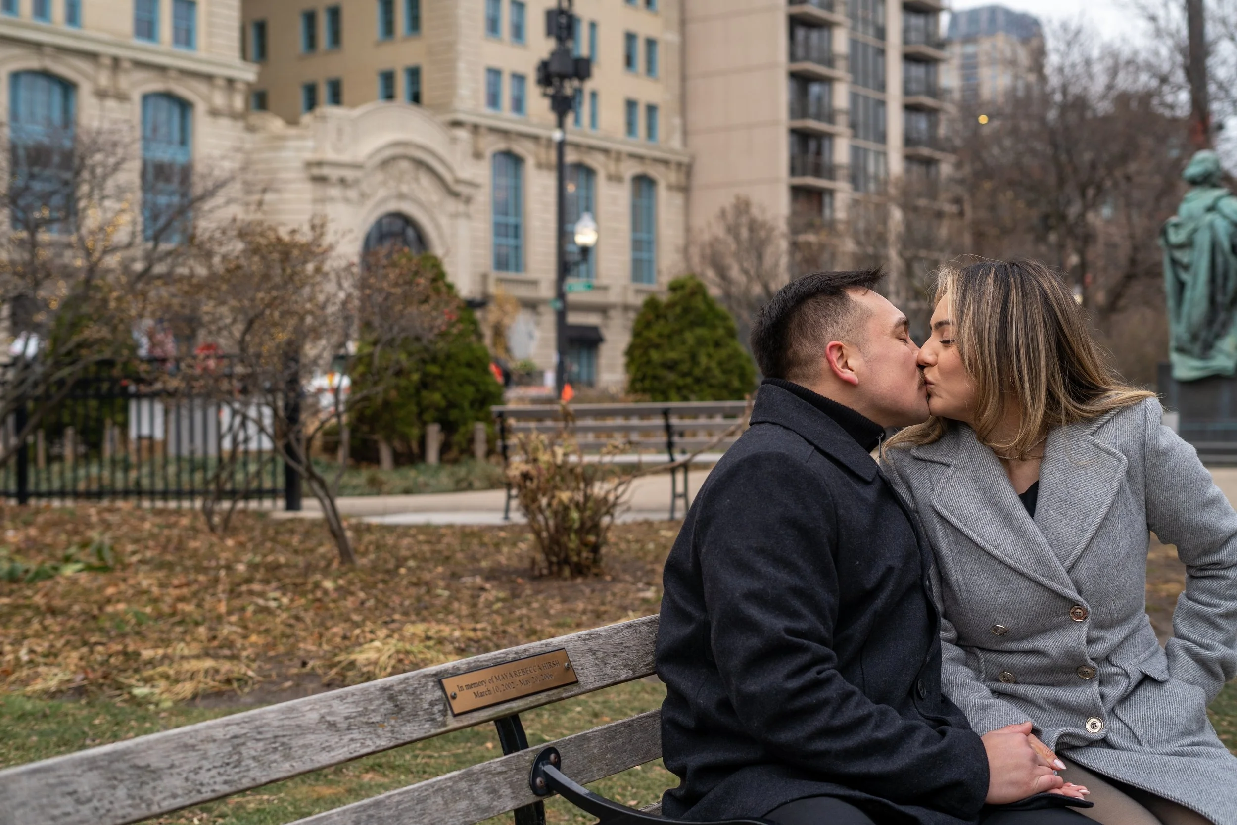 A man and woman sit closely on a park bench, sharing a kiss, with trees and buildings in the background.