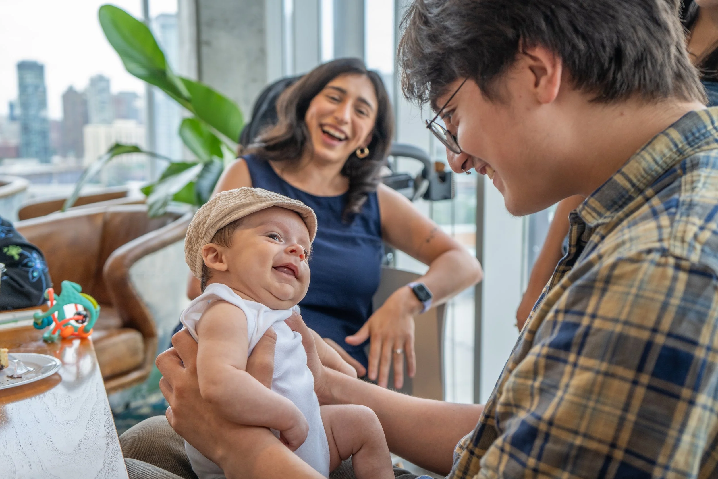 A man in glasses and a plaid shirt is holding a smiling baby wearing a sun hat, with a woman in a blue top laughing in the background. They are inside a room with large windows and city buildings outside.