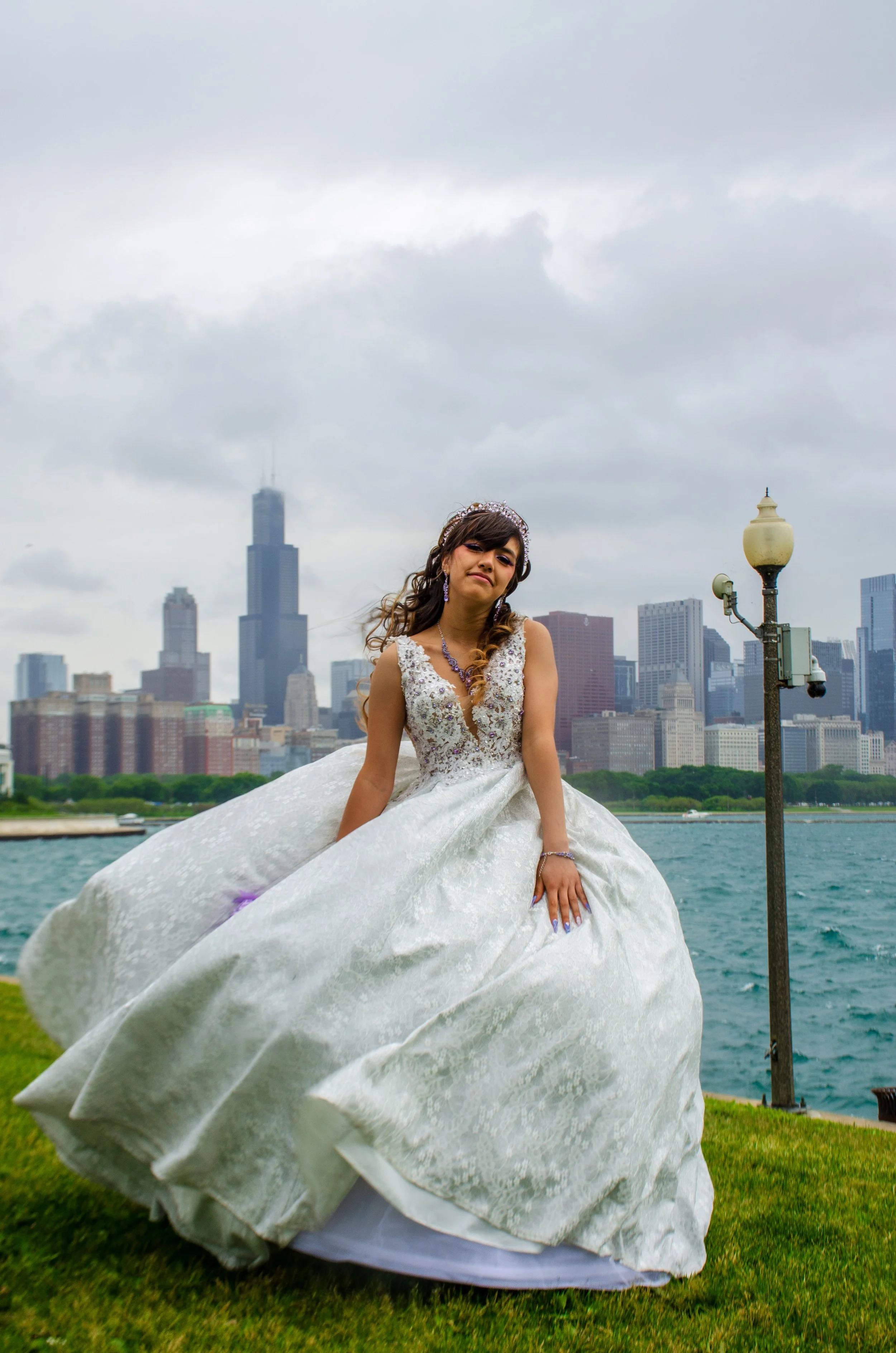 A young woman dressed in a white wedding gown standing outdoors near a river with a city skyline in the background and a gray sky overhead.