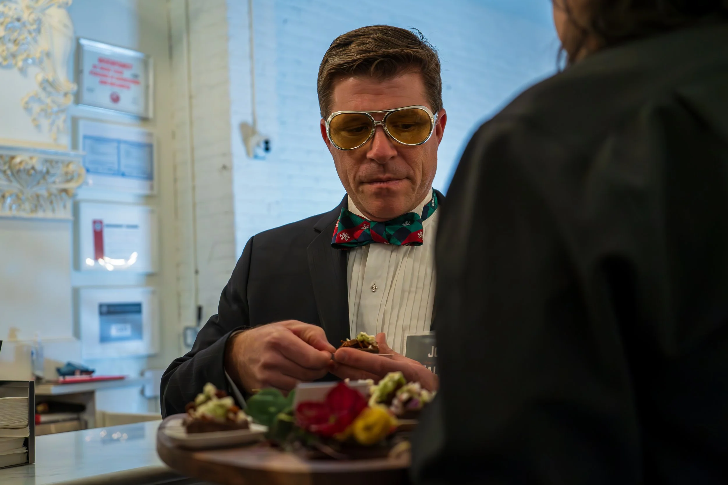 A man wearing a tuxedo, yellow-tinted sunglasses, and a holiday-themed bow tie, is serving food to a person with dark hair at a banquet or event. The background shows a white brick wall and decorative wall elements.