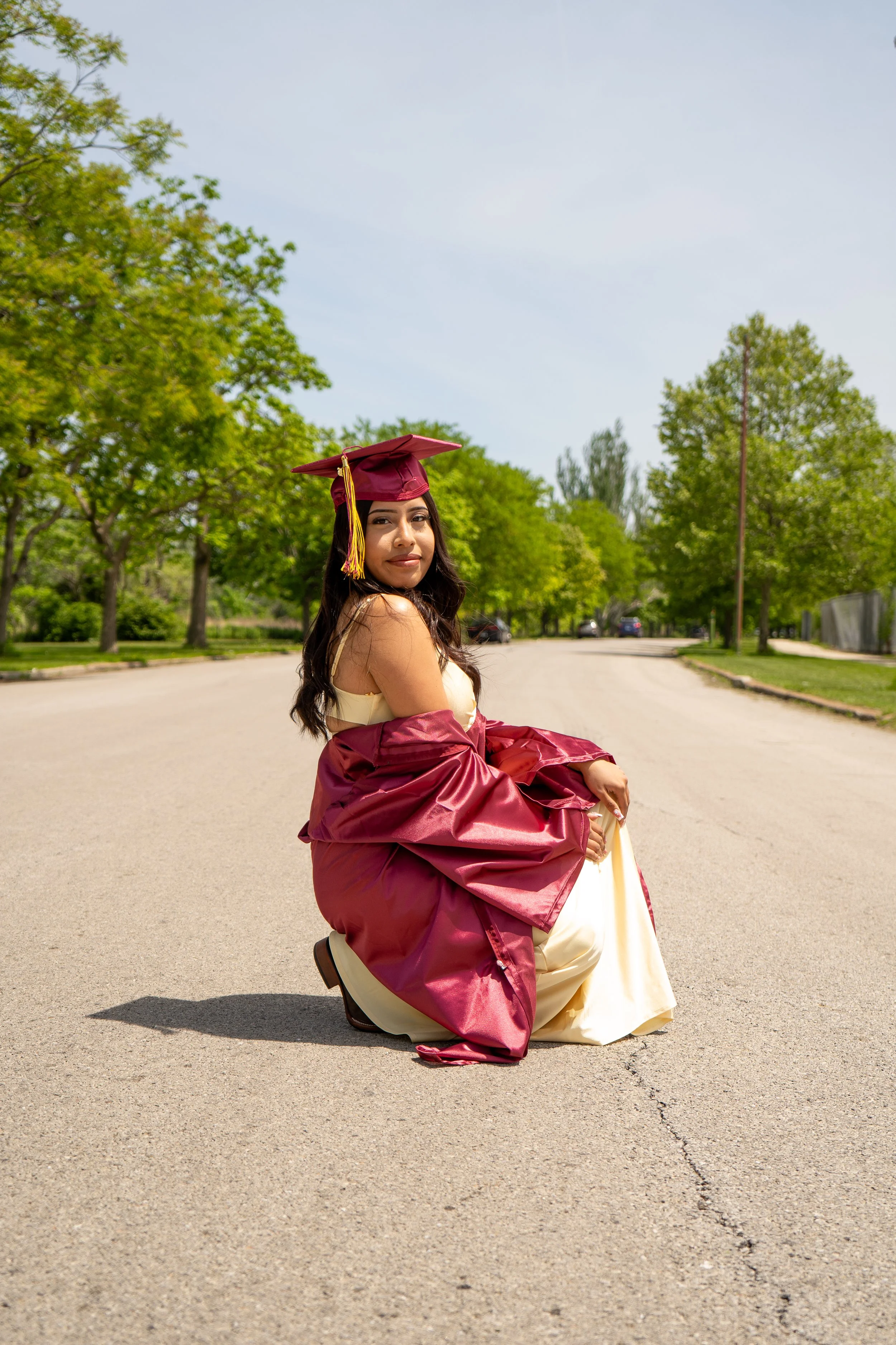 A young woman in a maroon graduation cap and gown kneeling on the street, surrounded by green trees and parked cars, with a blue sky overhead.