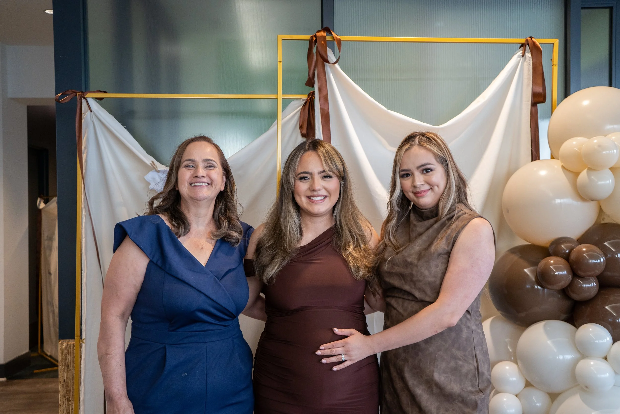 Three women standing together at an event, smiling at the camera, with a backdrop of balloons and fabric decorations.