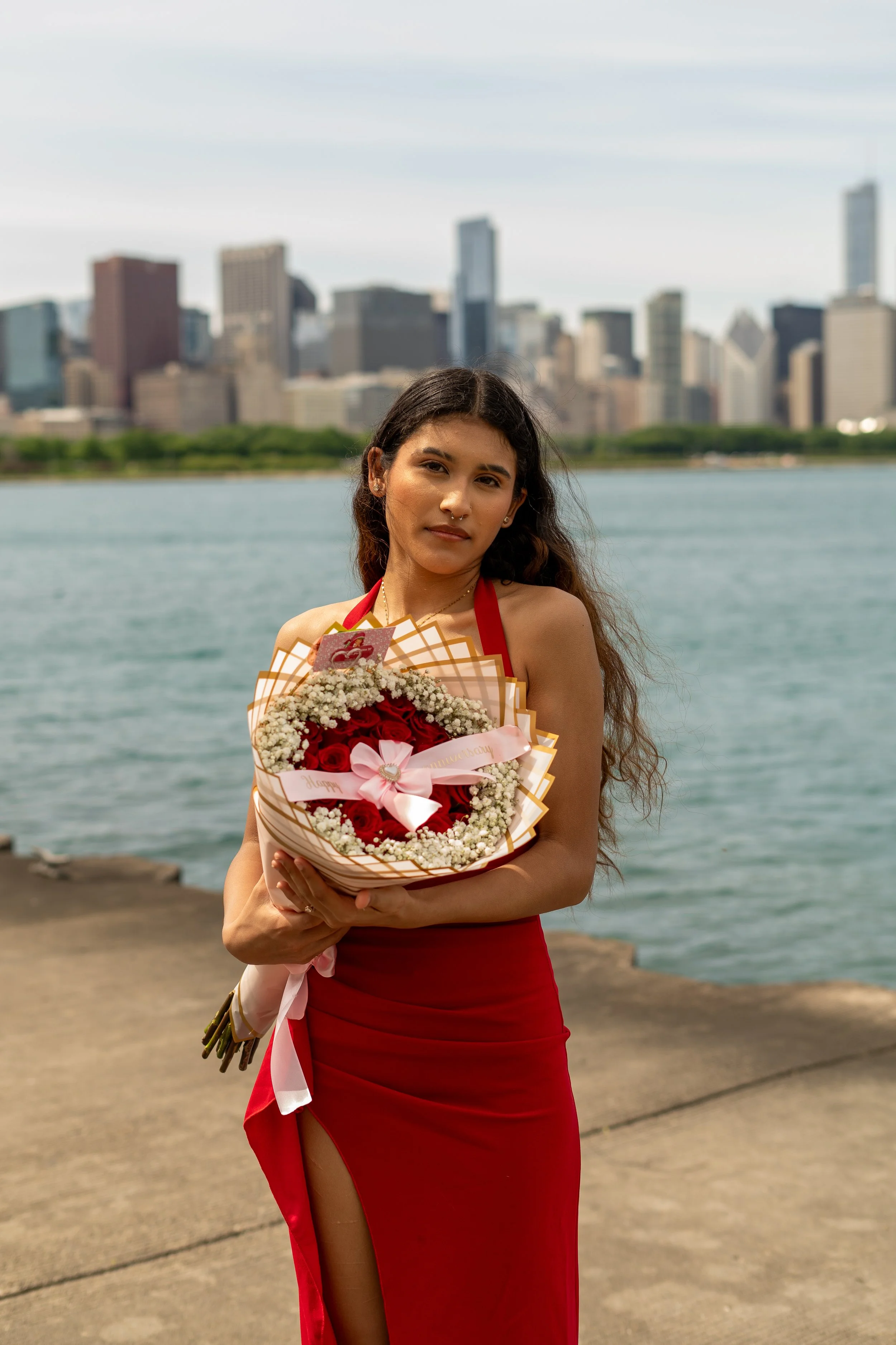 A woman in a red dress holding a bouquet of red and white flowers, standing outdoors near water with a city skyline in the background.