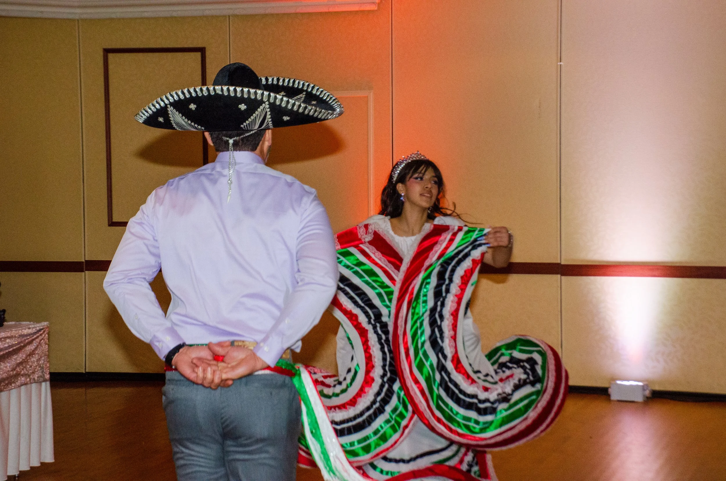 A man in a white shirt and a large traditional Mexican sombrero facing a woman. The woman is wearing a colorful, patterned dress and a tiara, and appears to be dancing.